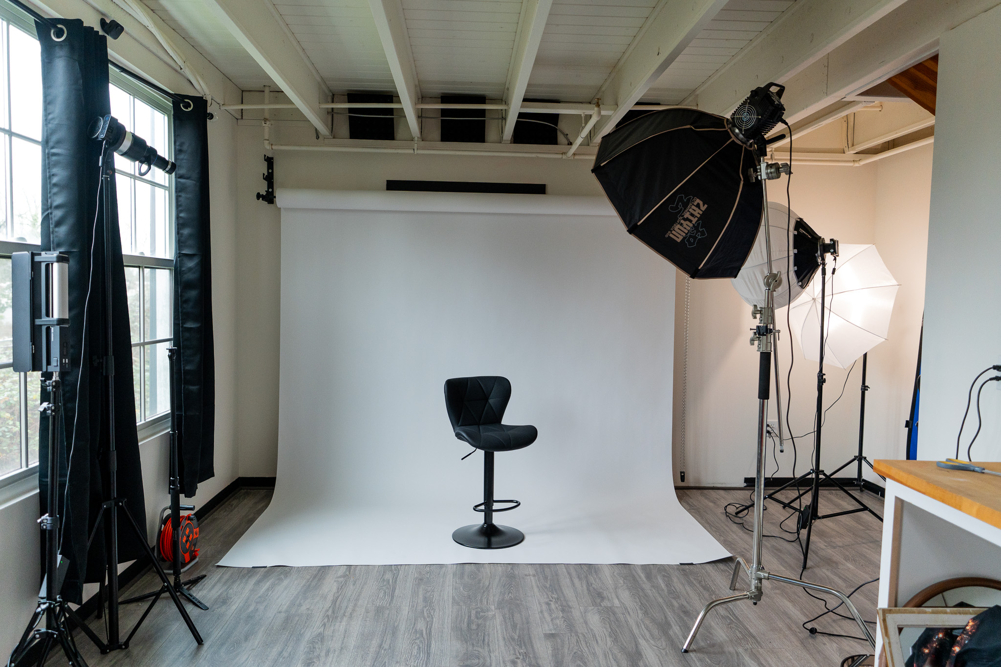 Photo of a photography studio with a black chair on a white backdrop, studio lights, and equipment, in a room with windows.