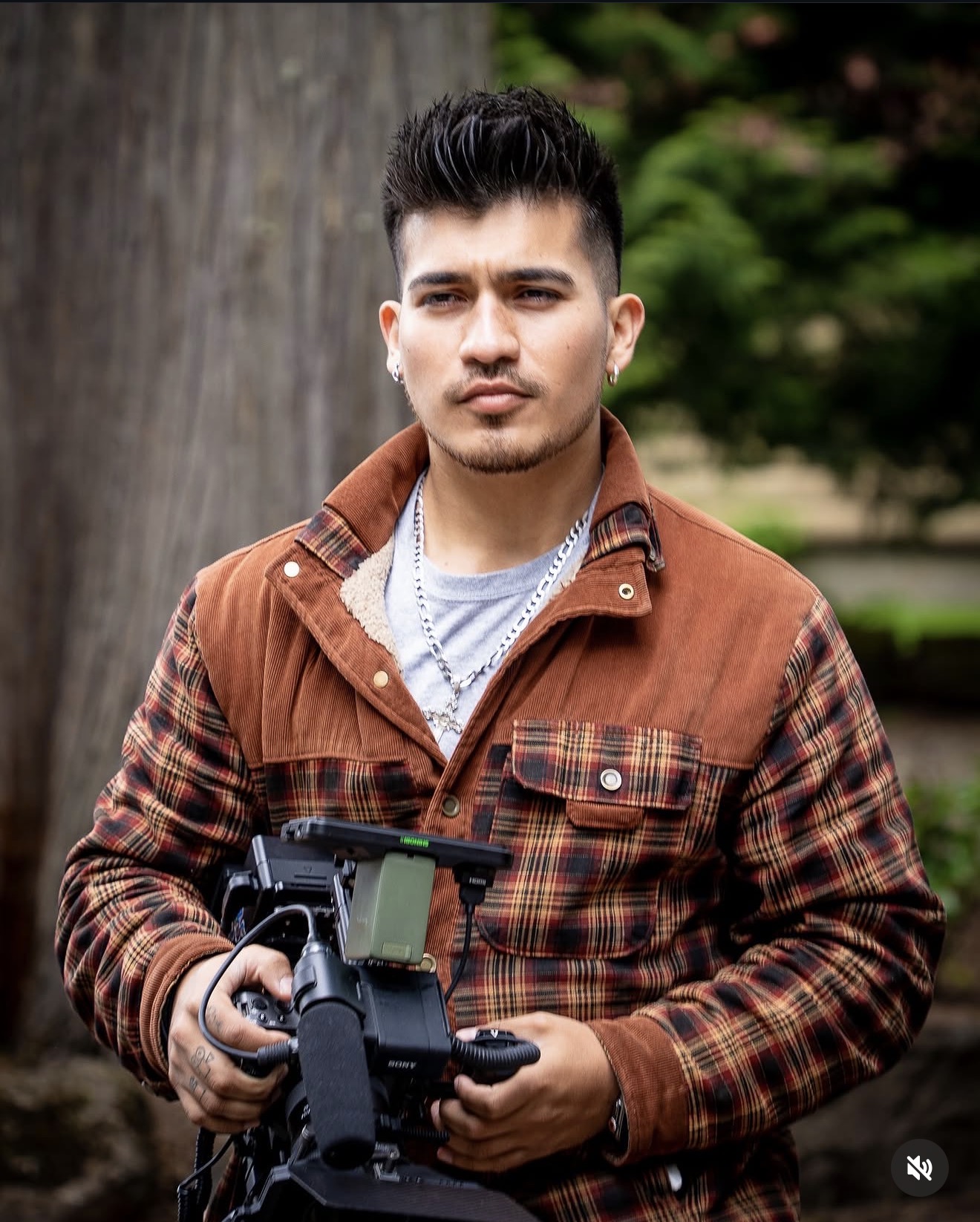 Young man with short dark hair and a mustache, wearing a brown plaid jacket, holding a camera outdoors.