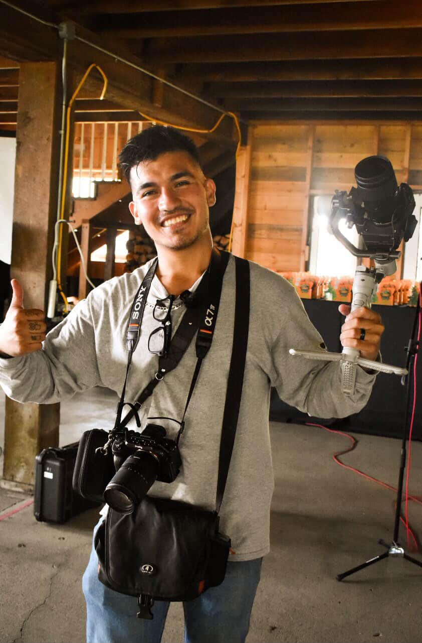 Young man smiling, holding a camera and a light, with a camera around his neck, in a wooden interior space.