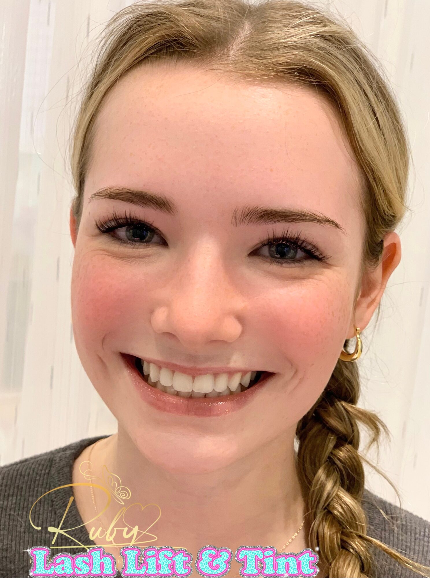 Young woman with blonde hair in a braid, smiling, wearing earrings and a dark top, indoors with light background.