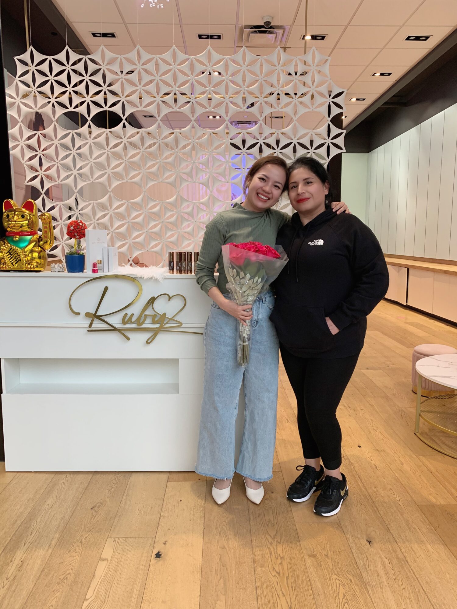 Two women standing together indoors, one holding a bouquet of flowers, smiling, with a decorative wall and counter behind them.
