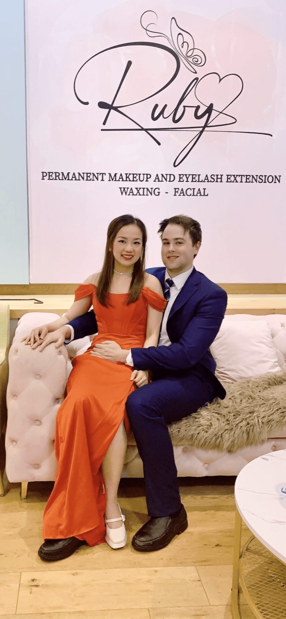 A woman in an orange dress and a man in a suit sit on a sofa, smiling, in a room with a pink sign behind them.