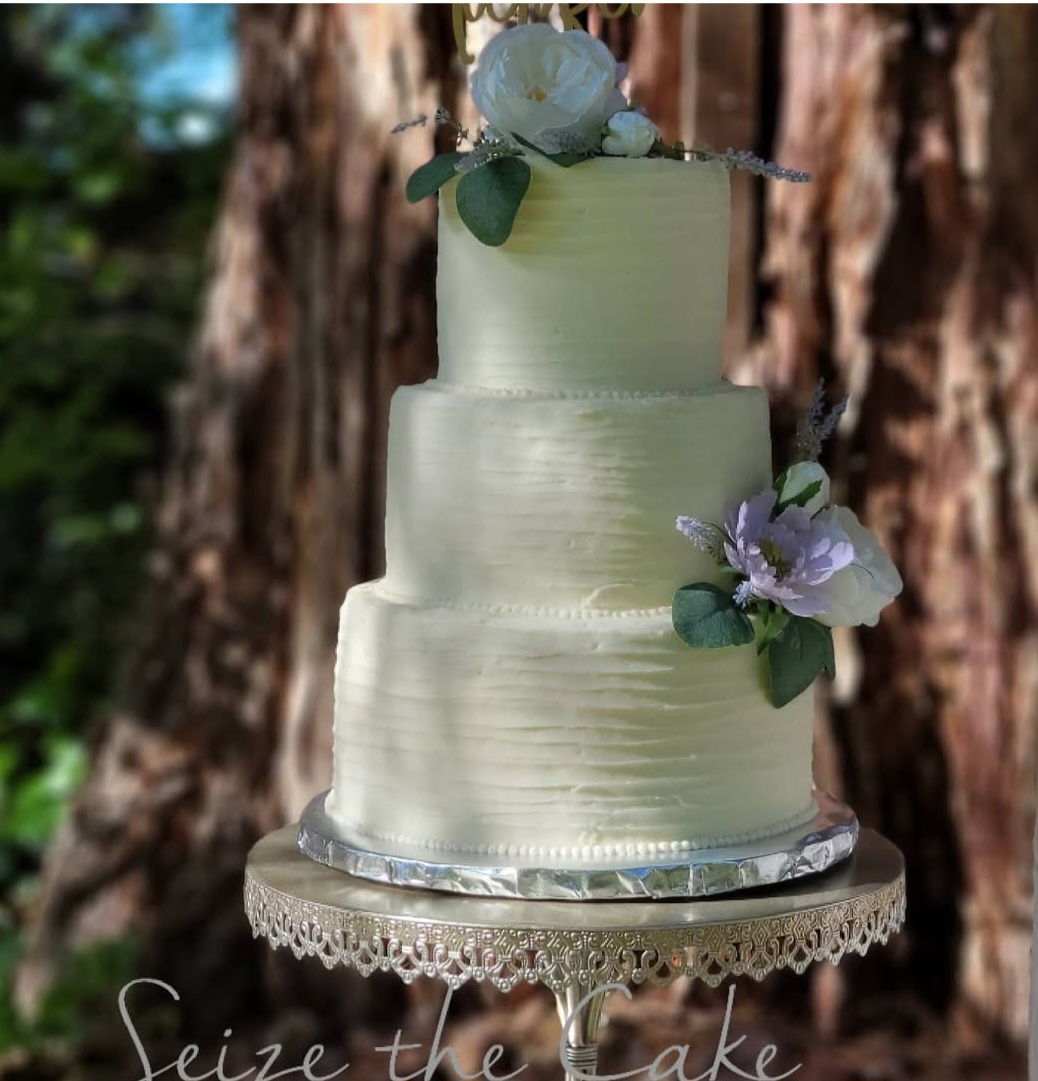 Three-tiered wedding cake with white frosting, decorated with flowers, on a decorative stand outdoors with trees in background.