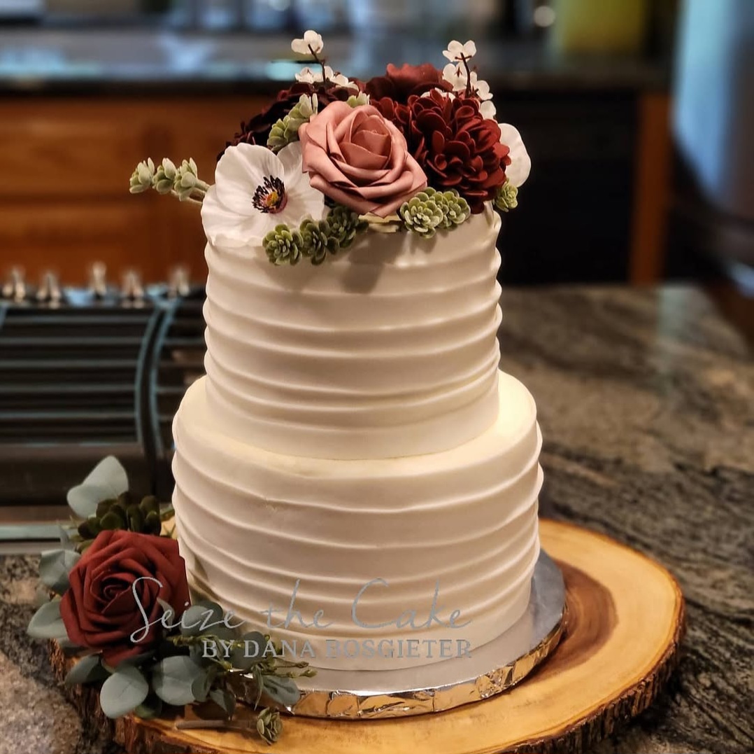 Two-tiered white cake with horizontal ridges, decorated with pink and red flowers on top and a flower arrangement at the base, on a wooden slab.