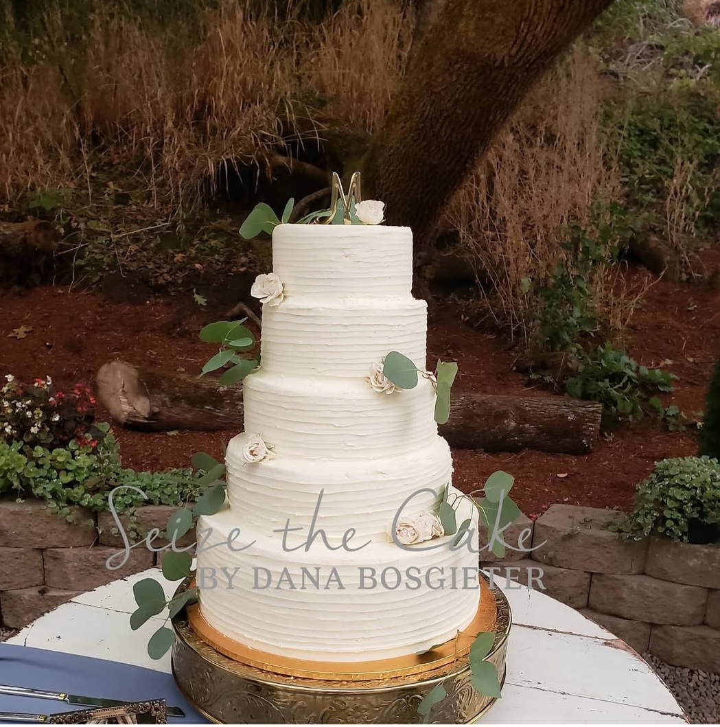 Three-tiered white wedding cake with green leaves and white flowers, on a wooden table outdoors, with trees and plants in background.