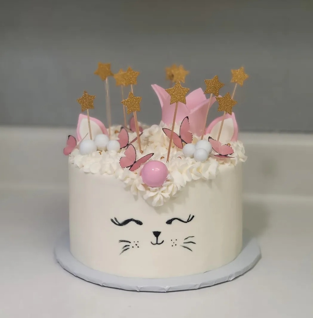Cake decorated with pink butterflies, gold stars, and white cream, featuring a cat face with closed eyes and whiskers.