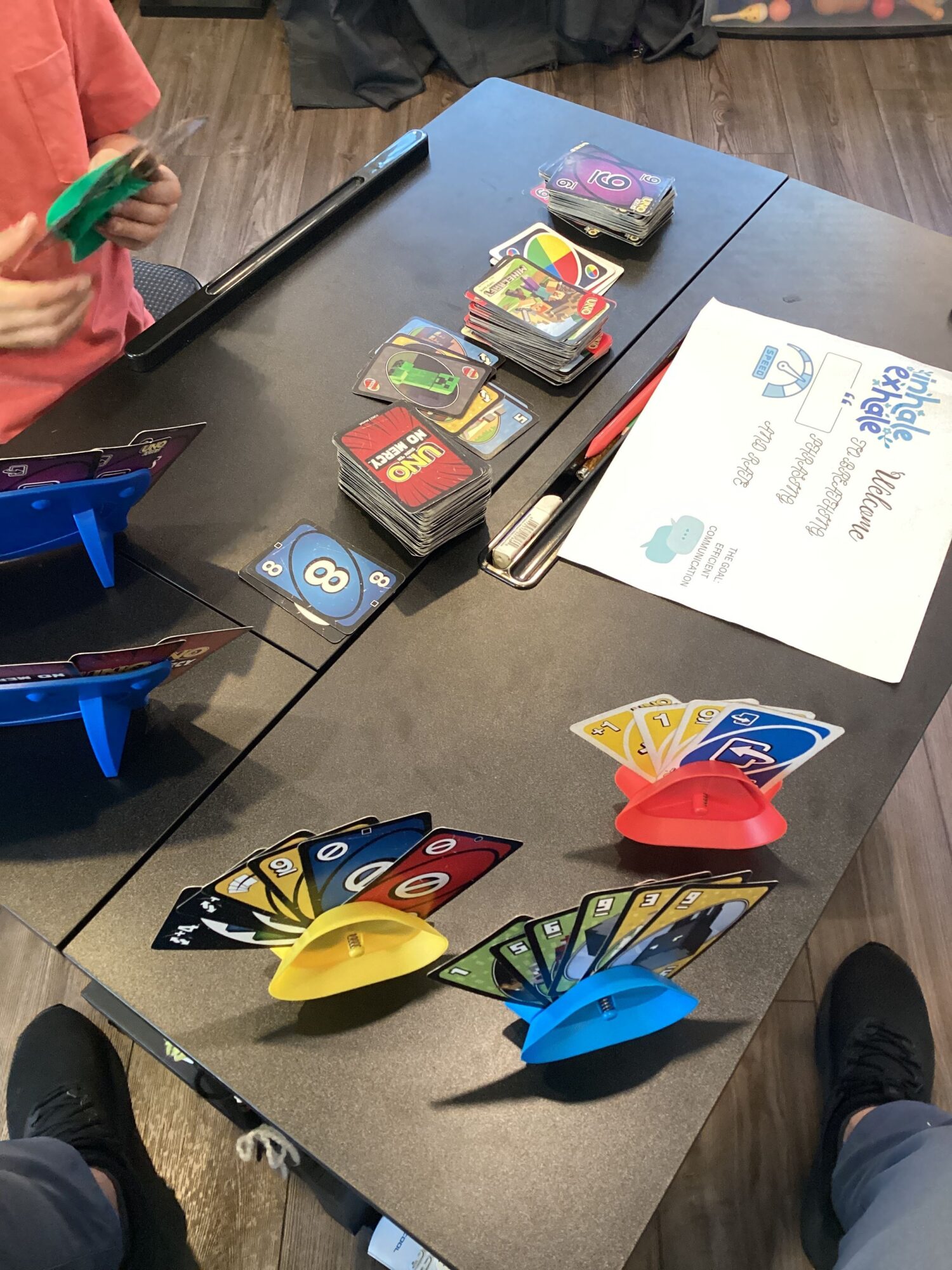 Table with colorful cards, paper, and game pieces, with people standing around it, in a room with wooden flooring.