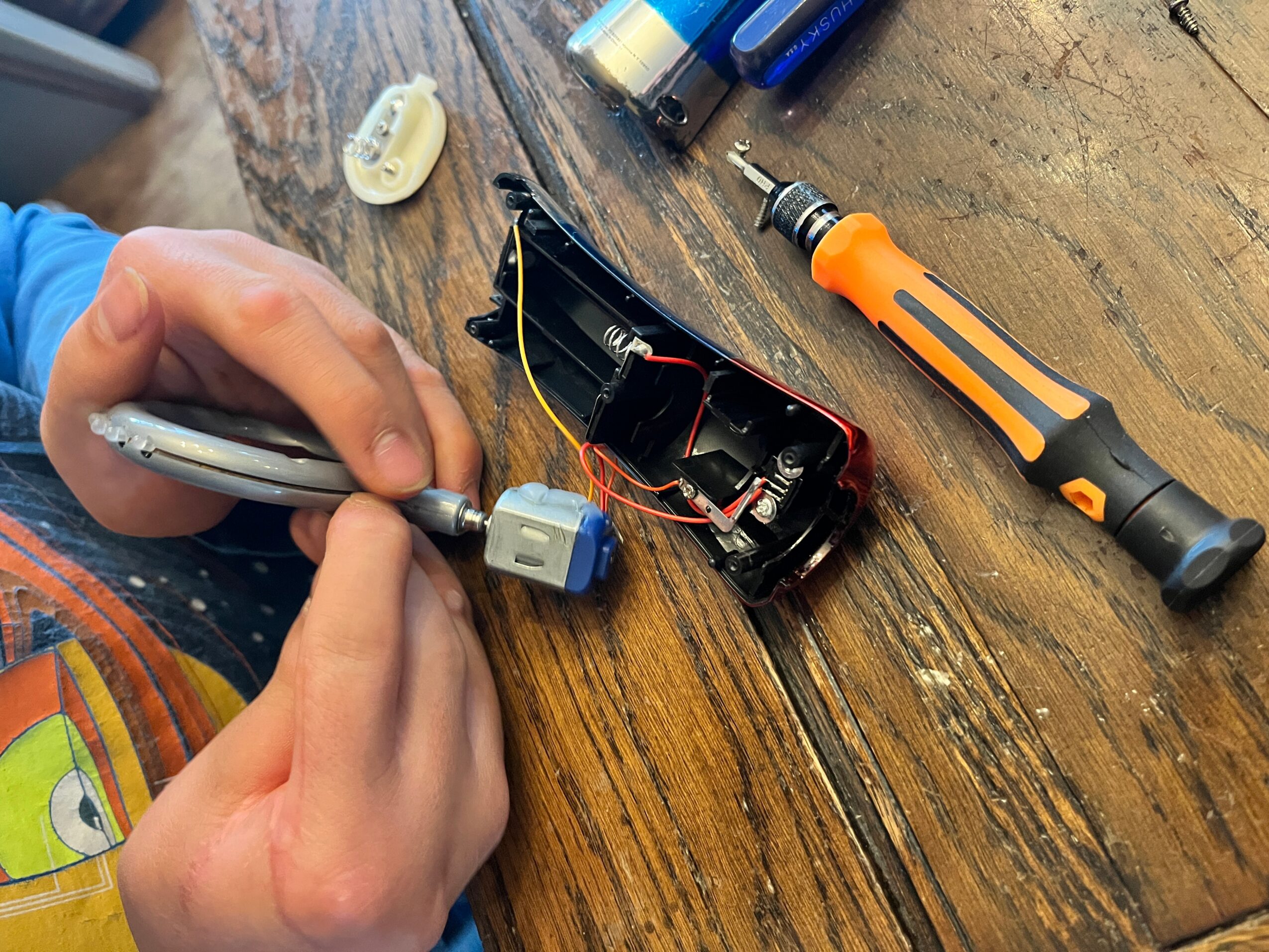 Person working on a small electronic device with tools on a wooden surface, including a screwdriver and a plastic component.