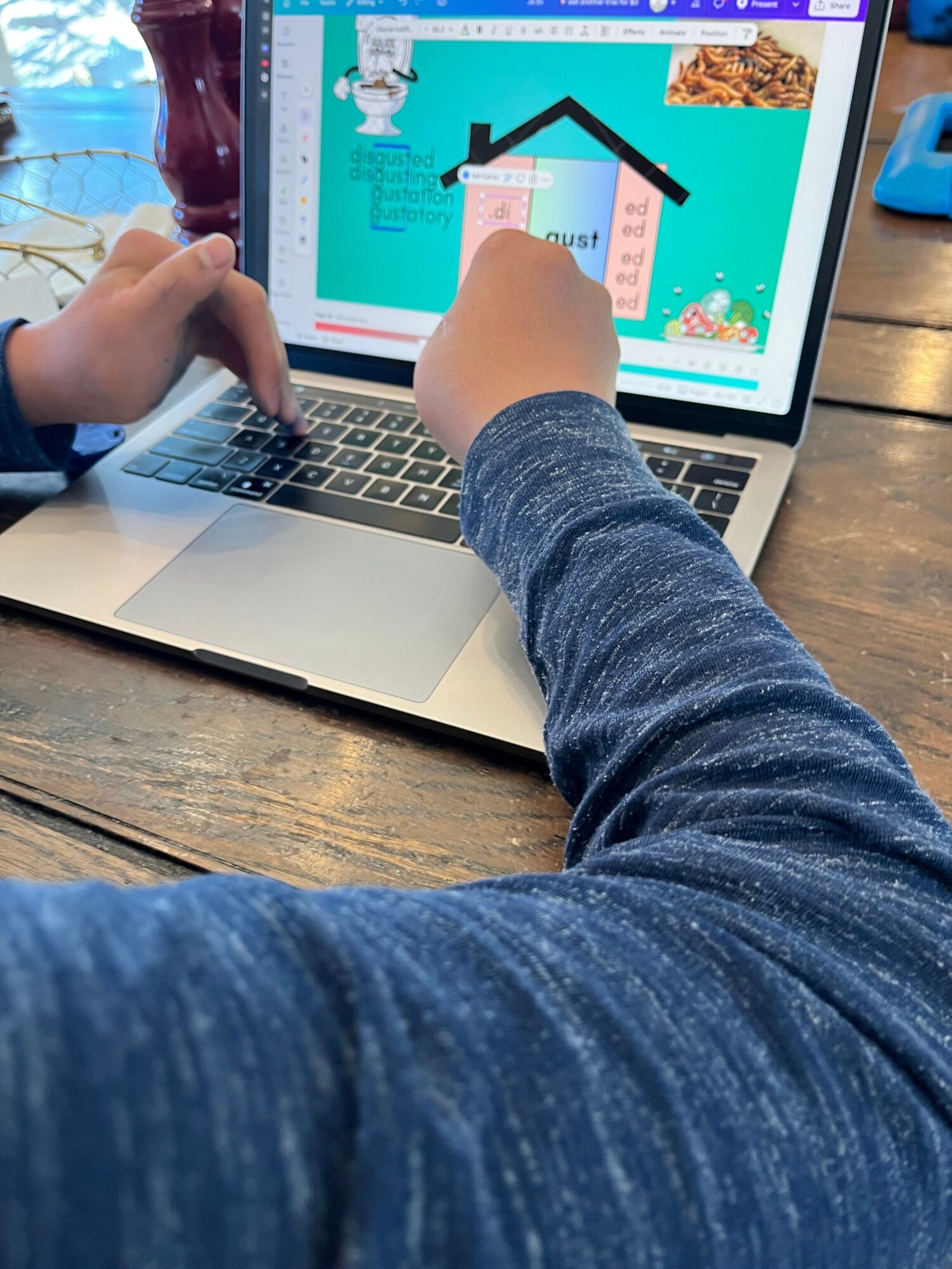 Child's hand points at a colorful game on a laptop screen, with another hand on the keyboard, on a wooden table.