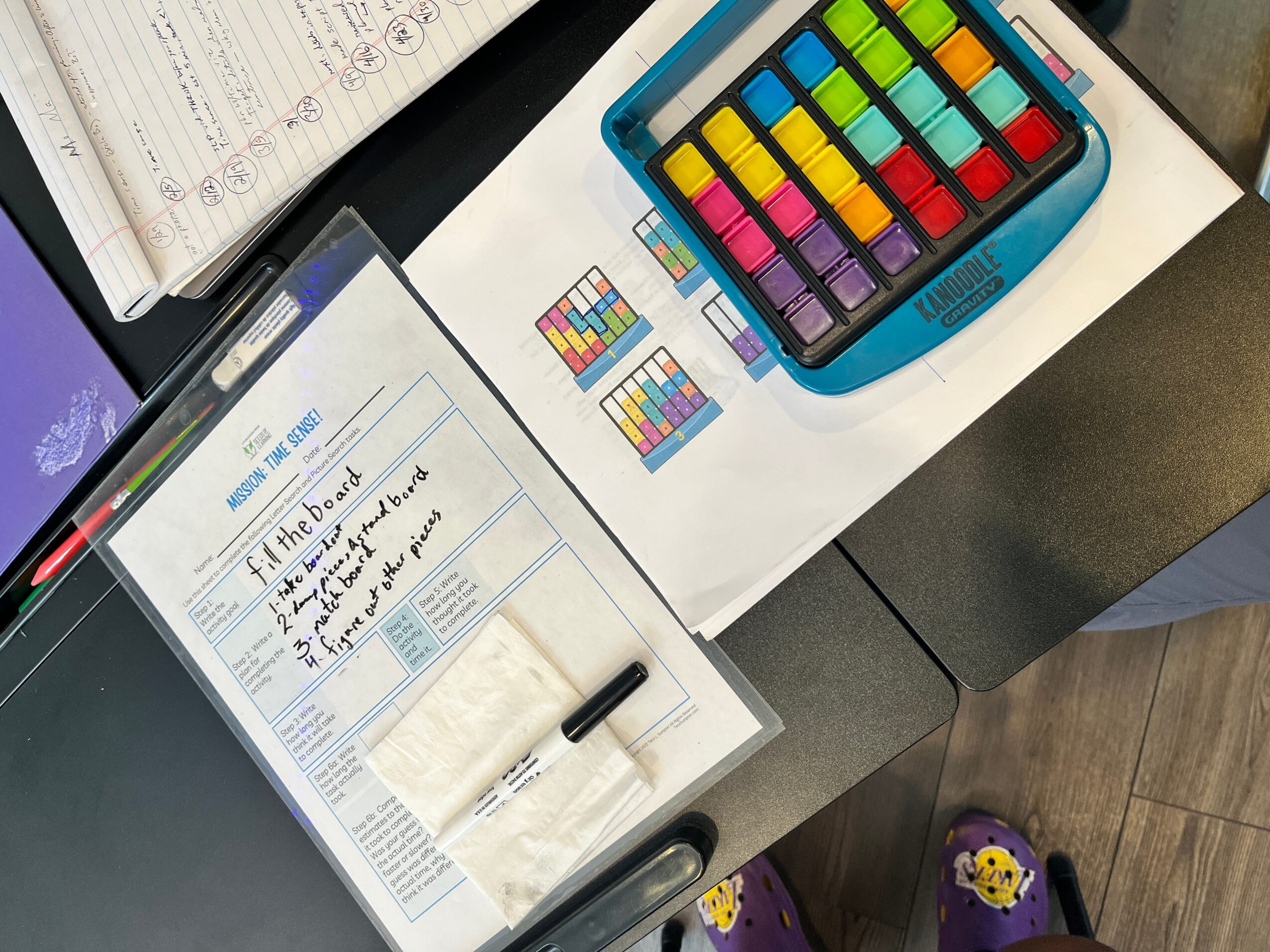 Open notebook with handwritten notes, colorful chalk set in a blue case, and small color samples on paper on a desk.