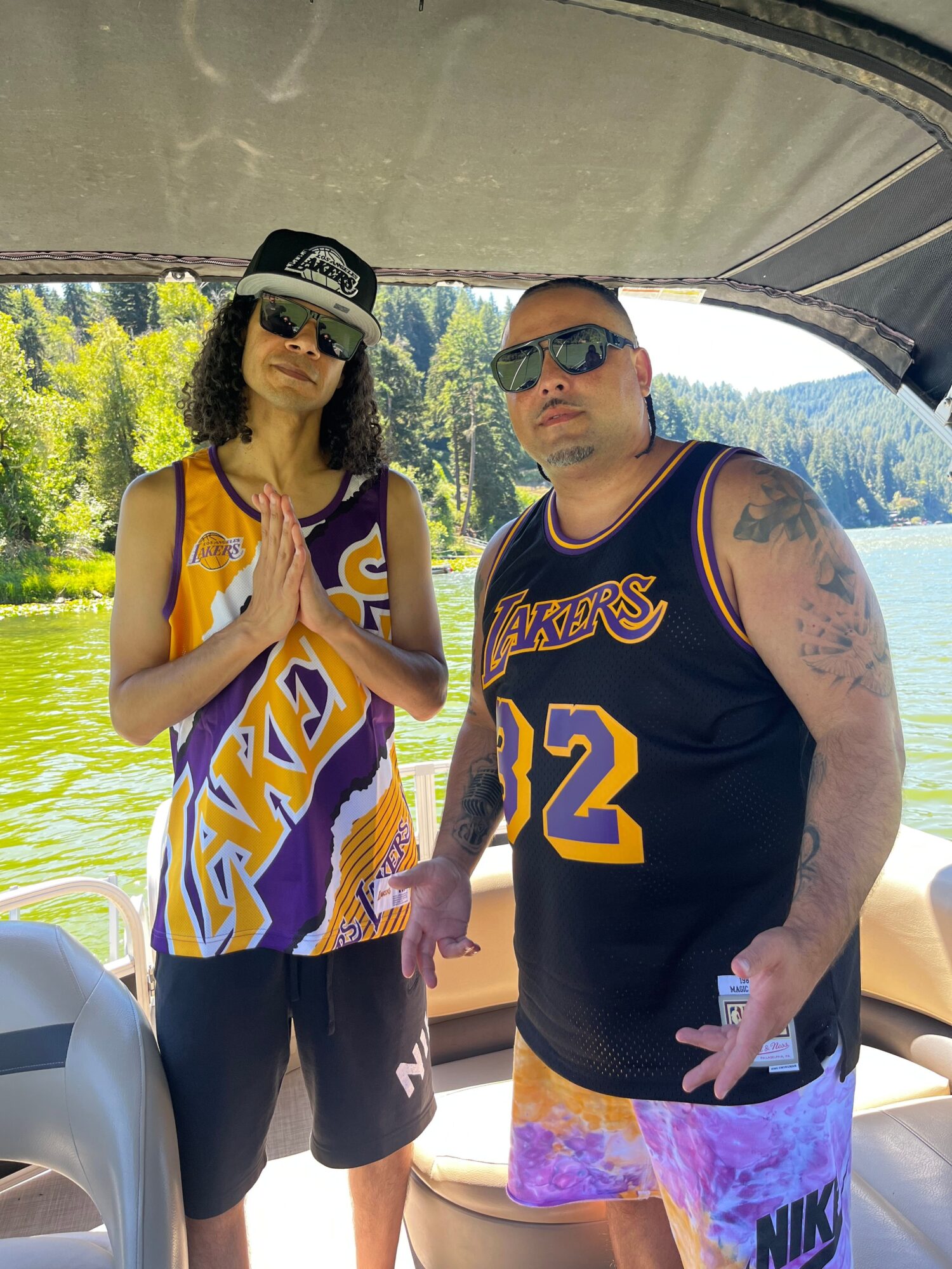 Two people standing outdoors near water, one wearing sunglasses and a Lakers jersey, the other in a Lakers tank top and cap, both smiling.