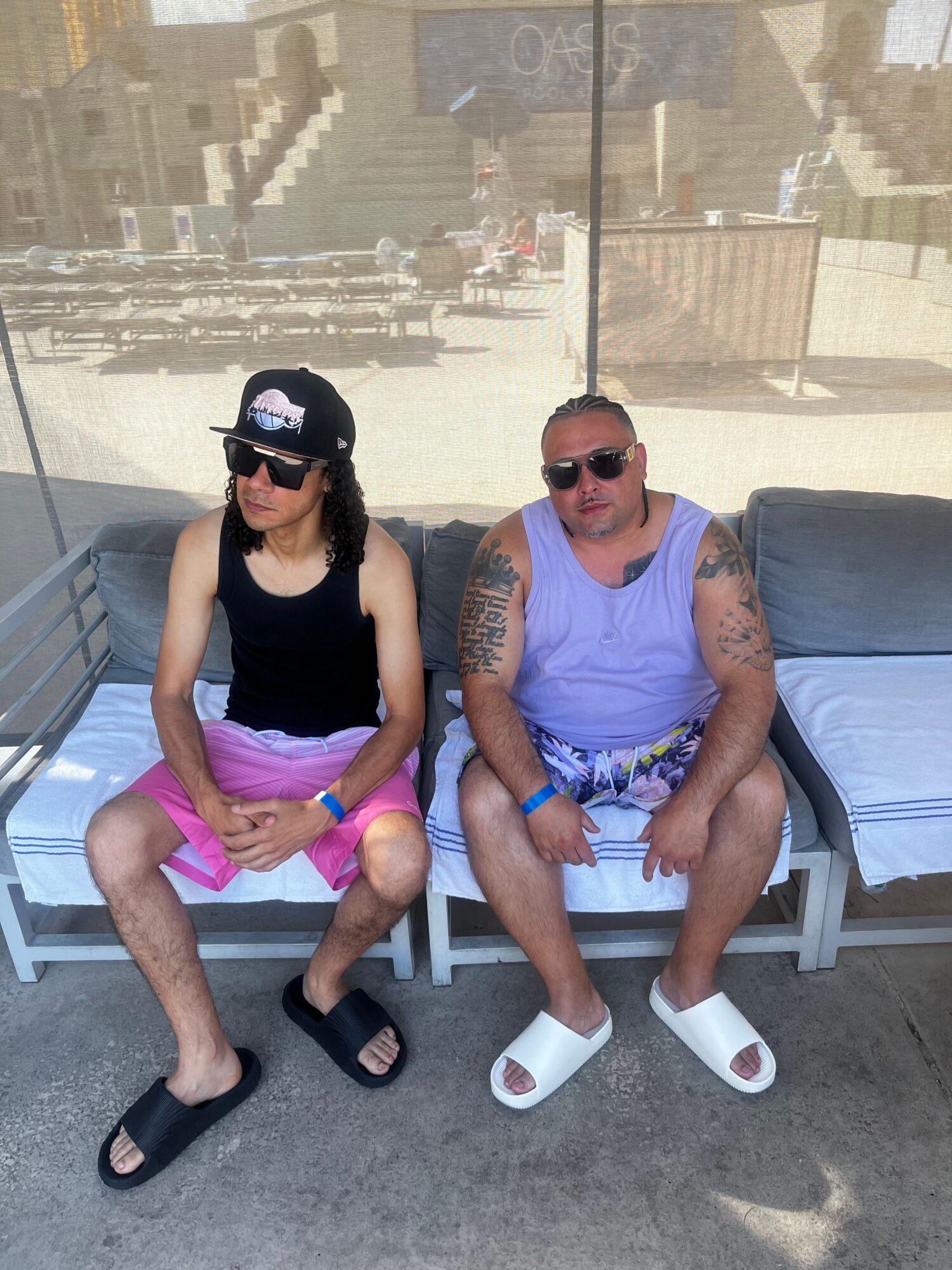Person sitting on a bench with tattoos, wearing sunglasses, a purple shirt, and shorts, with a cityscape background.
