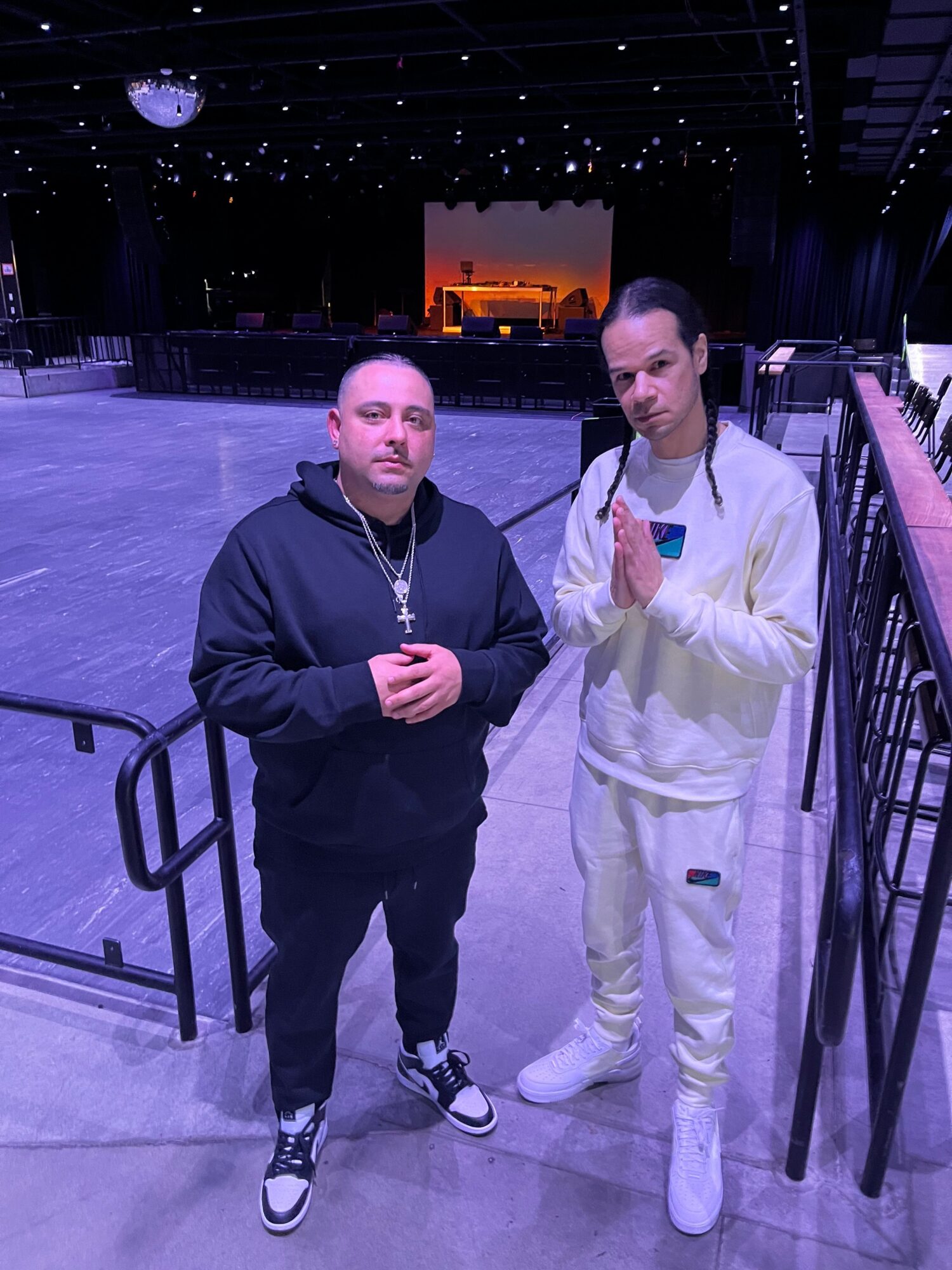 Two people standing indoors, one in white tracksuit and the other in black, with a railing and dark background behind them.
