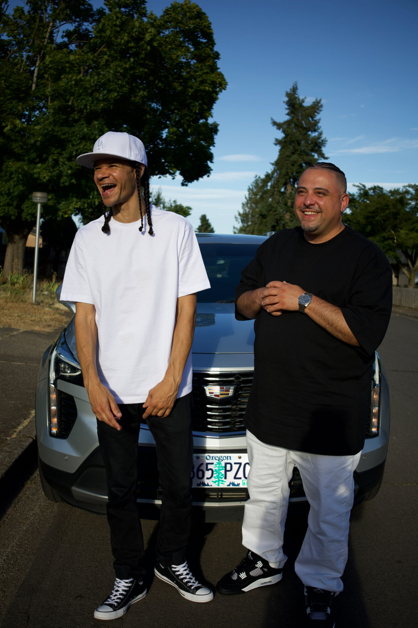 Two men standing outdoors in front of a car, smiling, with trees and a blue sky in the background.