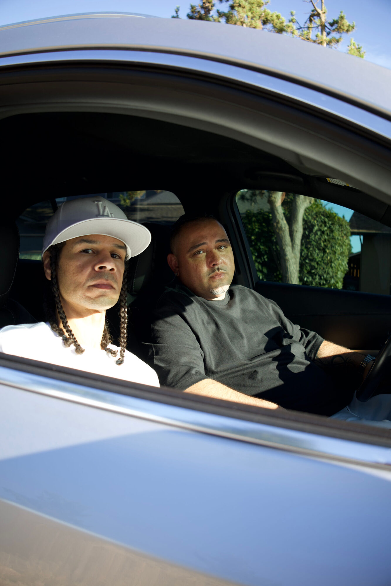 Two people sitting in a car, one wearing a white hat and the other with a shaved head, looking at the camera.
