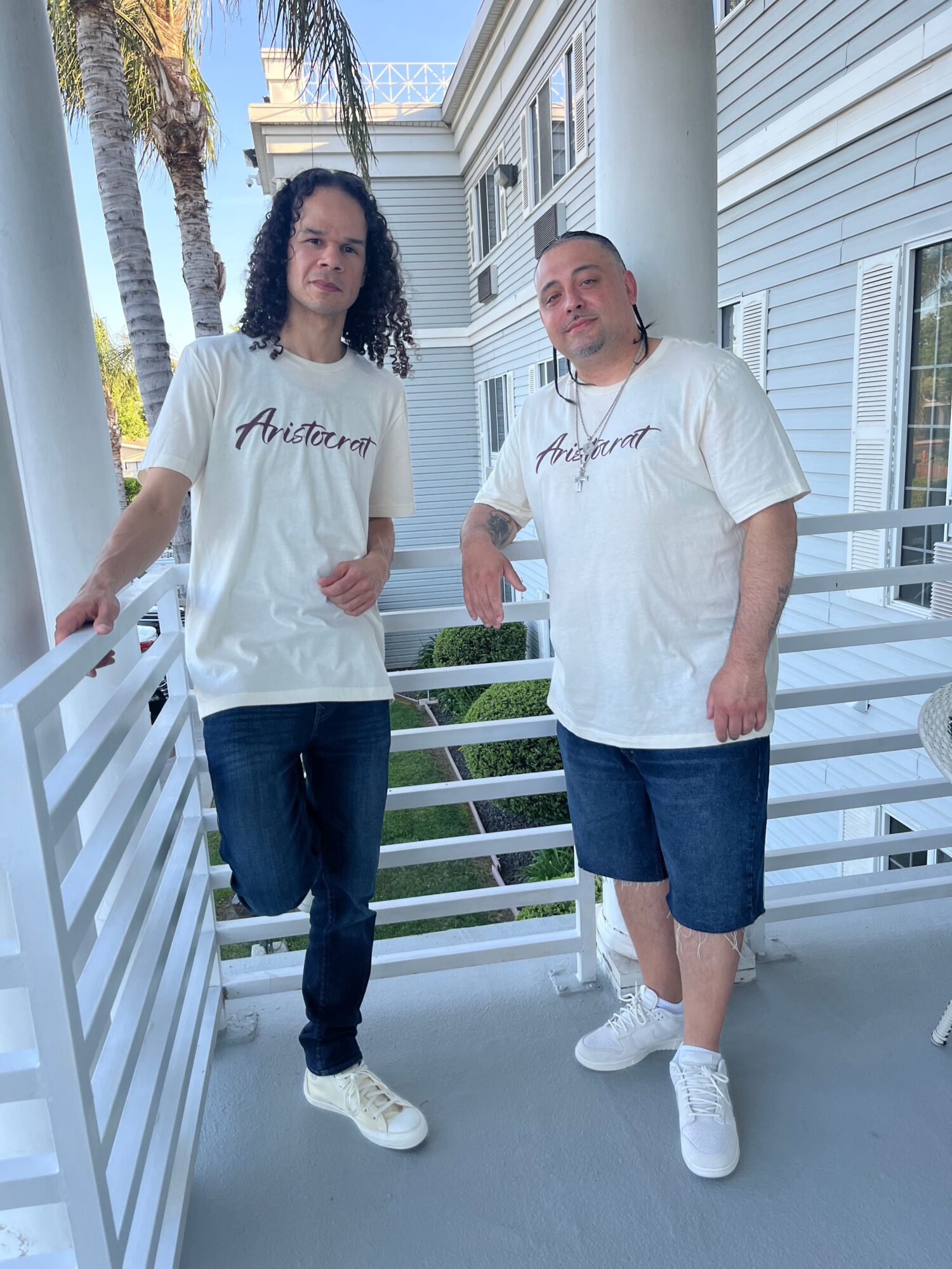 Two people standing on a balcony with white railings, wearing casual clothing, with a house and trees in the background.