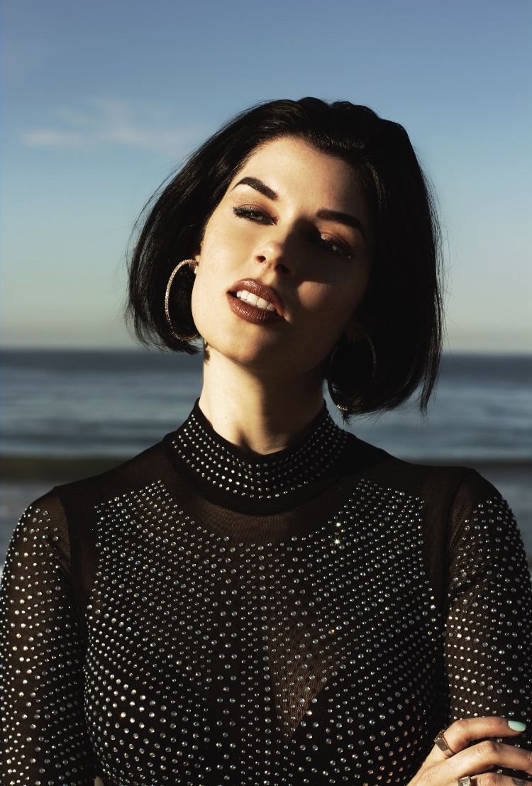Young woman with short black hair and hoop earrings stands outdoors near water, wearing a black, dotted, sheer top.