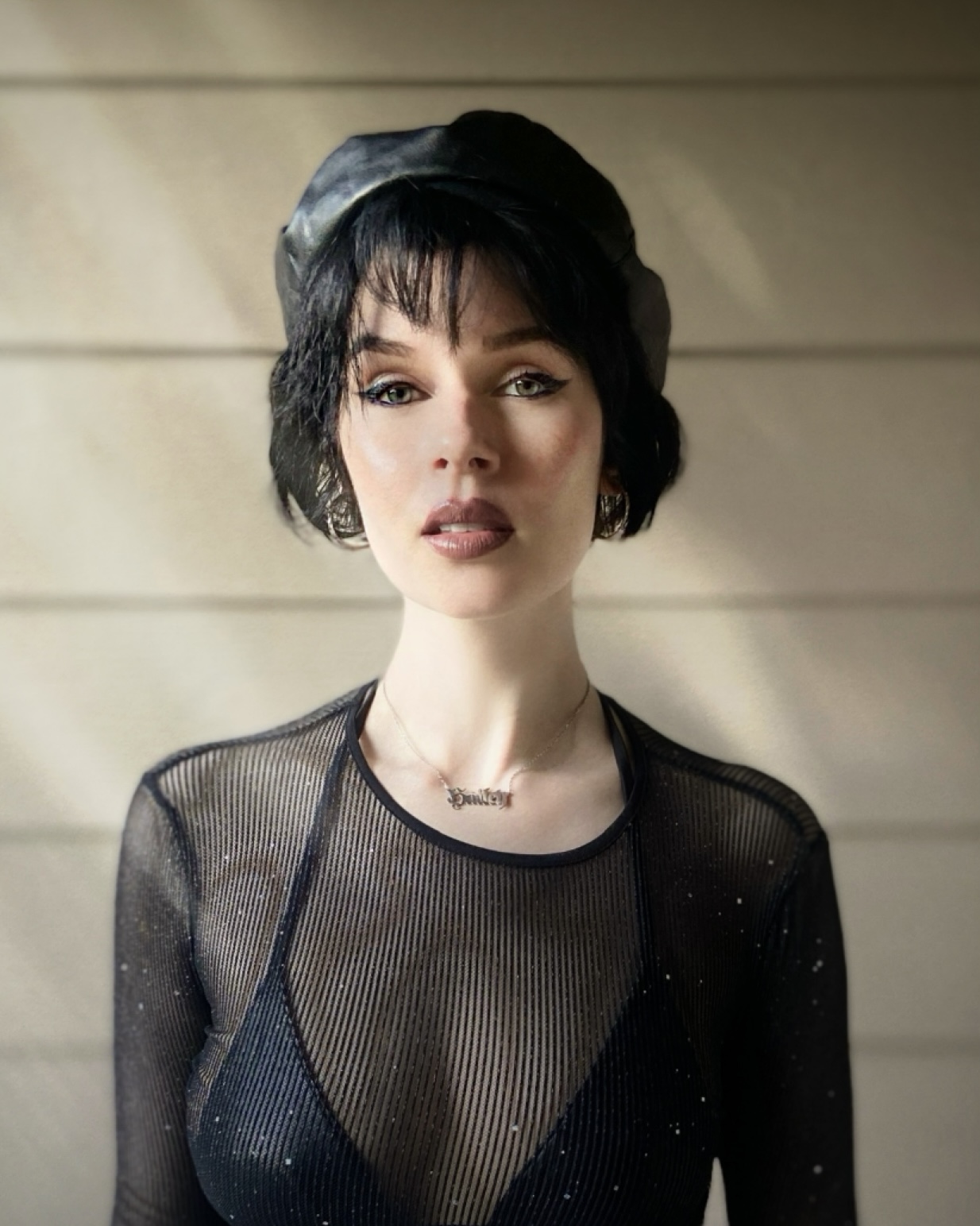 Portrait of a woman with short dark hair wearing a black mesh top and a black hat, standing against a light background.