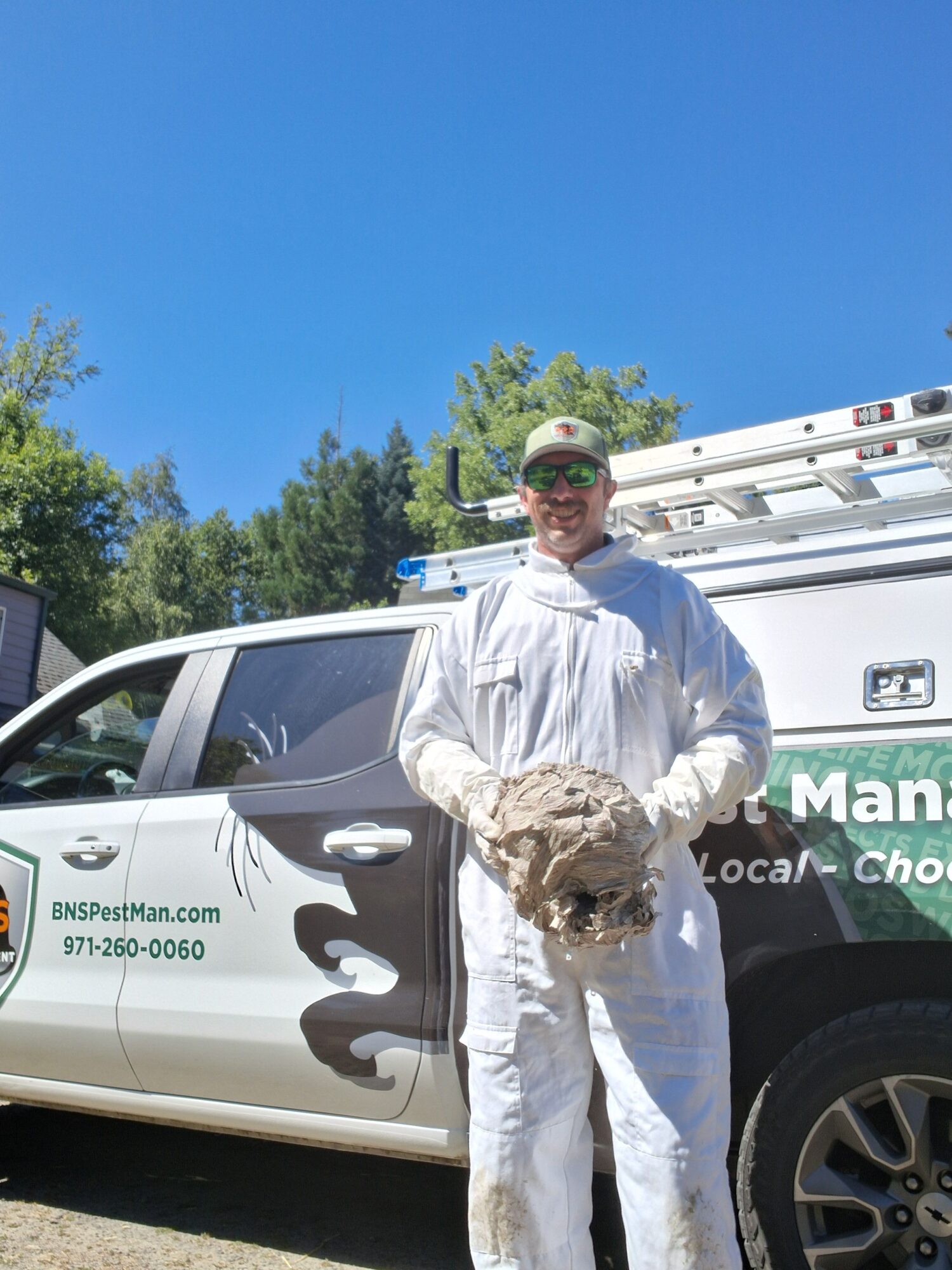Person in white protective suit holding a small dog, standing next to a white vehicle with blue sky and trees in background.