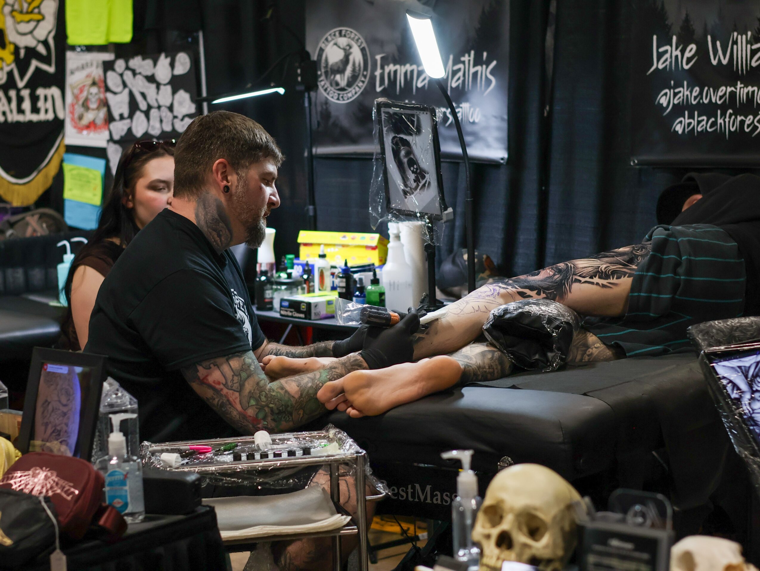 Tattoo artist working on a person's arm in a tattoo studio with various supplies and artwork visible.