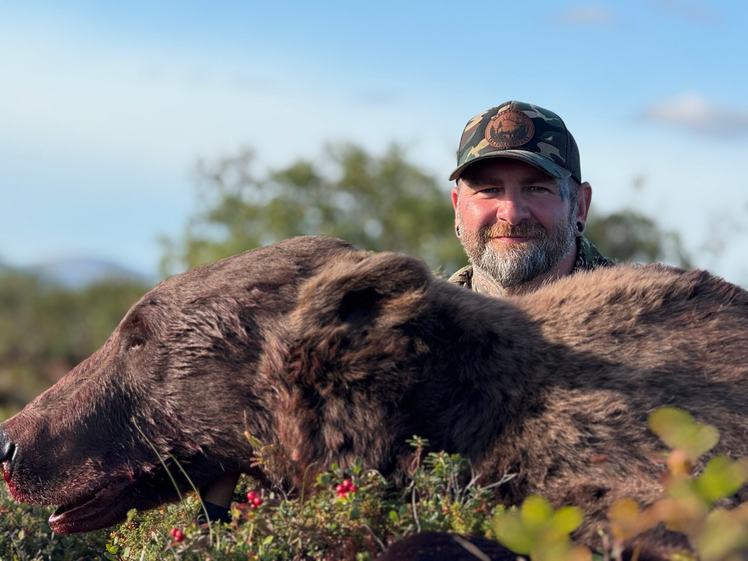 Man with a beard and camouflage cap smiling outdoors with two large brown animals, possibly bears, in front of him.