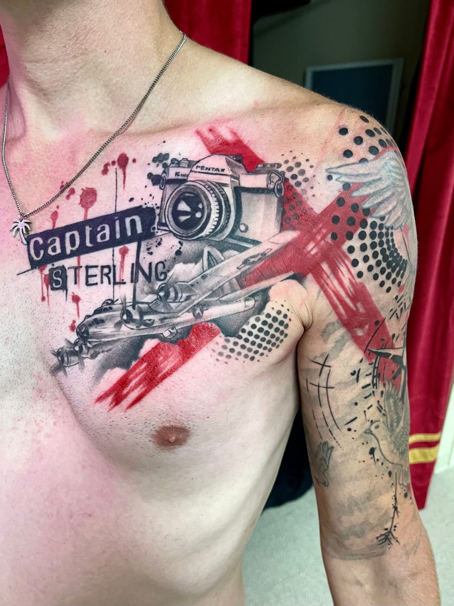 Tattoo of a camera, airplane, and text on a person's chest and shoulder with red and black ink splashes.