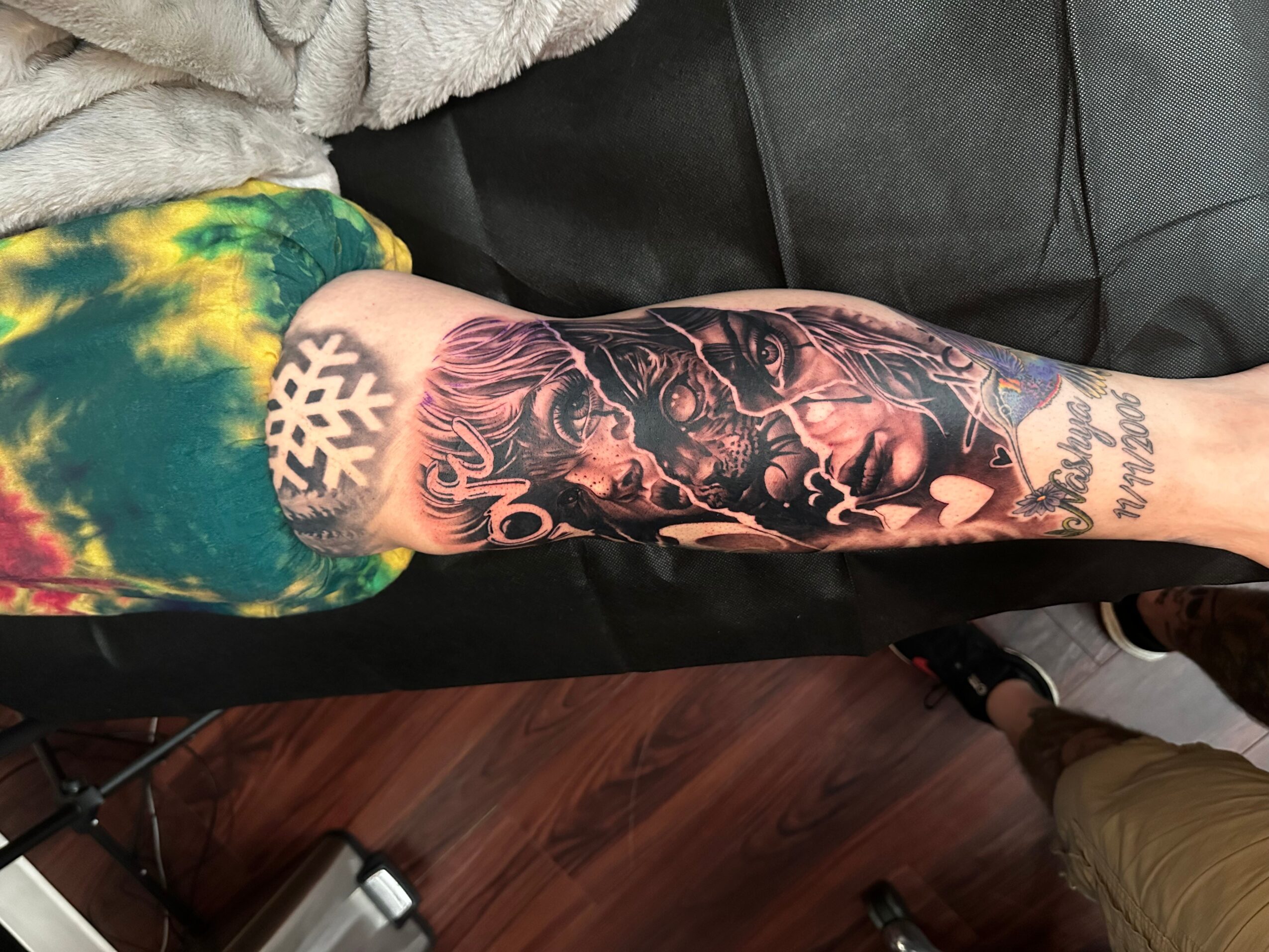 Person with a colorful tie-dye shirt and a large detailed tattoo on their arm, sitting on a black couch.