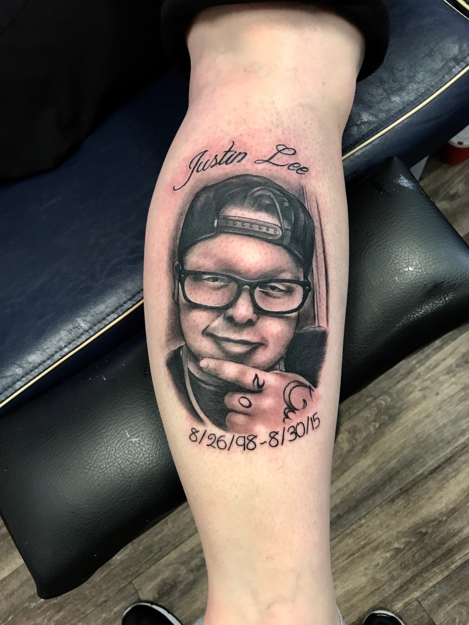 Tattoo of a young person with glasses and a cap, smiling, with a finger near their lips, and dates below.
