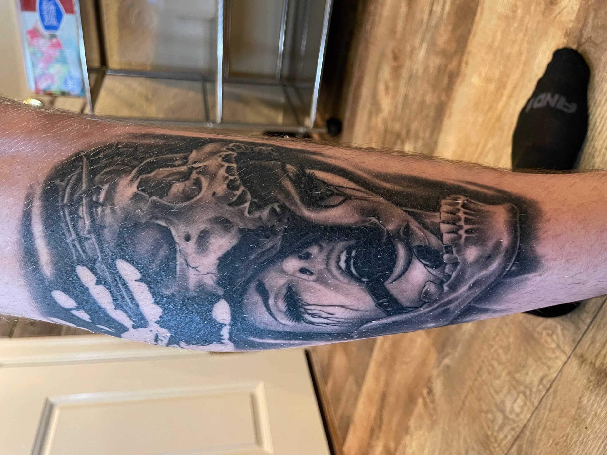 Black and gray tattoo of a skull, a woman, and abstract elements on a person's forearm.