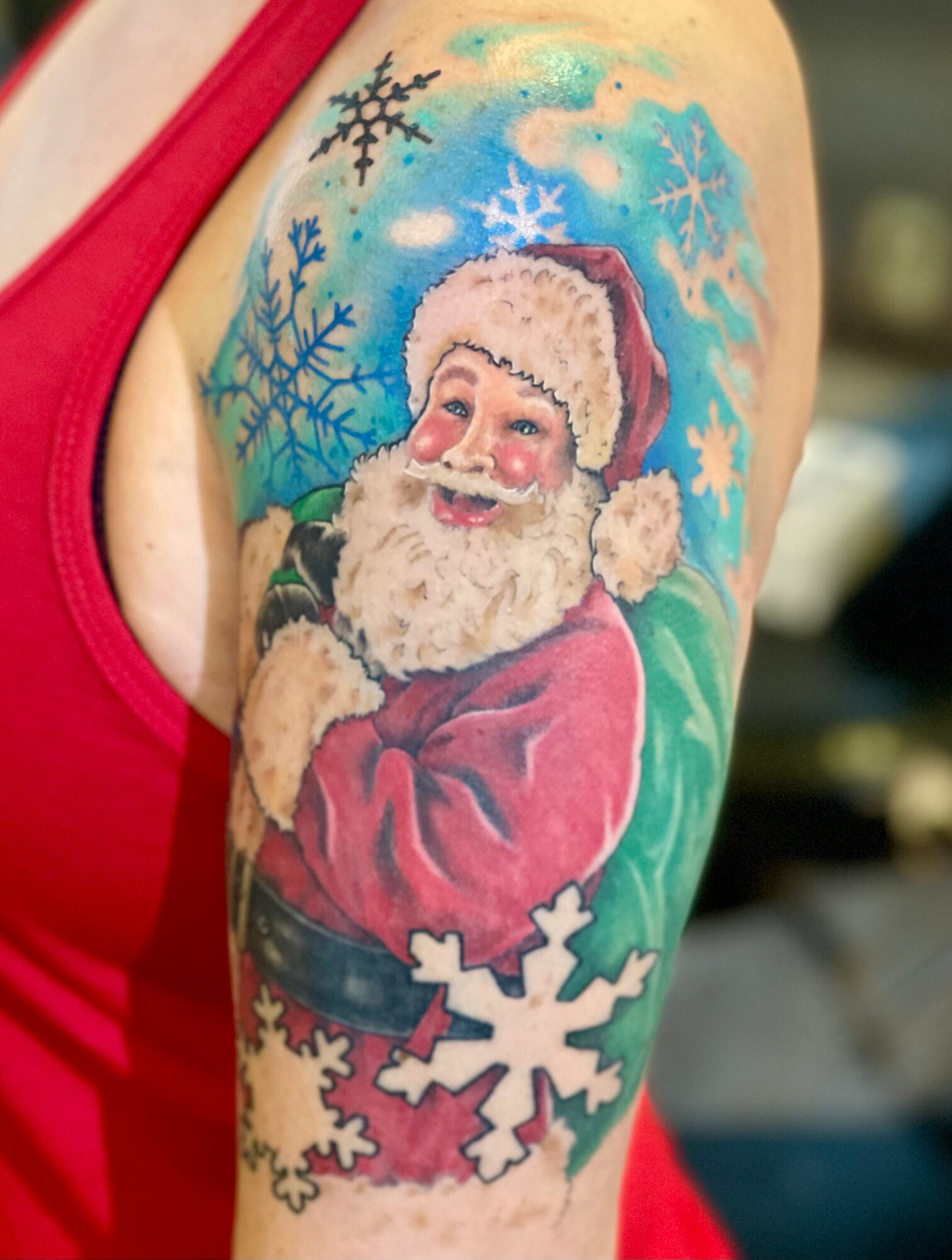 Colorful tattoo of Santa Claus with snowflakes on upper arm, background with blue sky and snowflakes, person wearing red tank top.