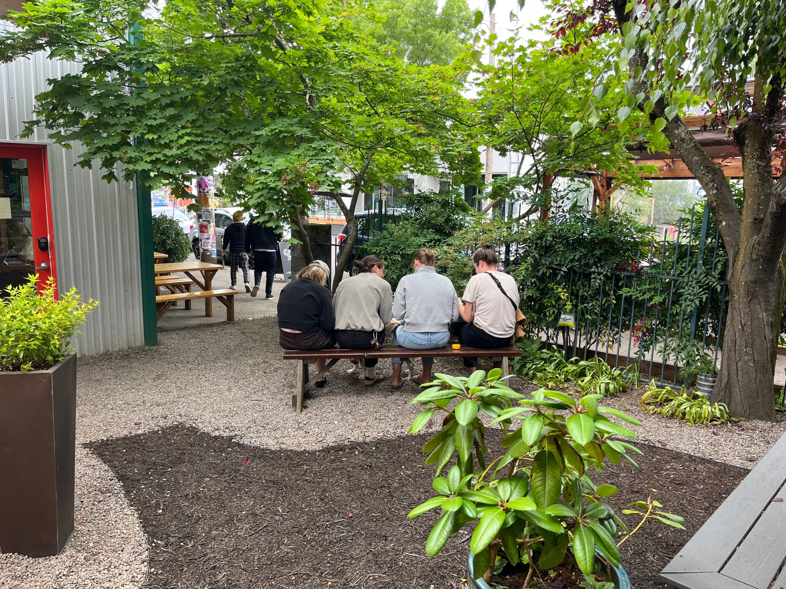 Four people sit on a bench outdoors under trees, with a garden and pathway nearby.