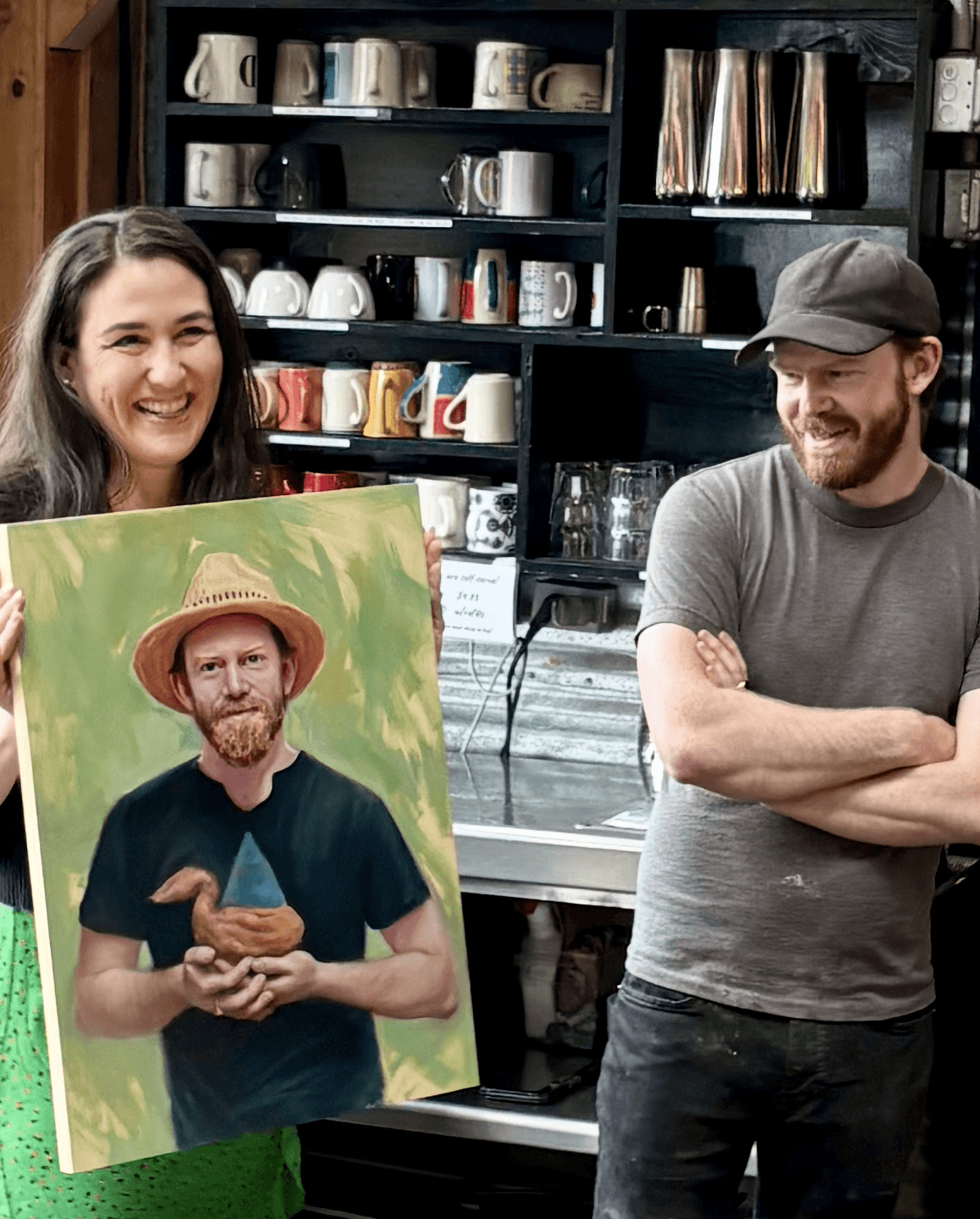 Two people smiling in a coffee shop, one holding a painting of a man with a hat and beard, shelves with cups behind them.