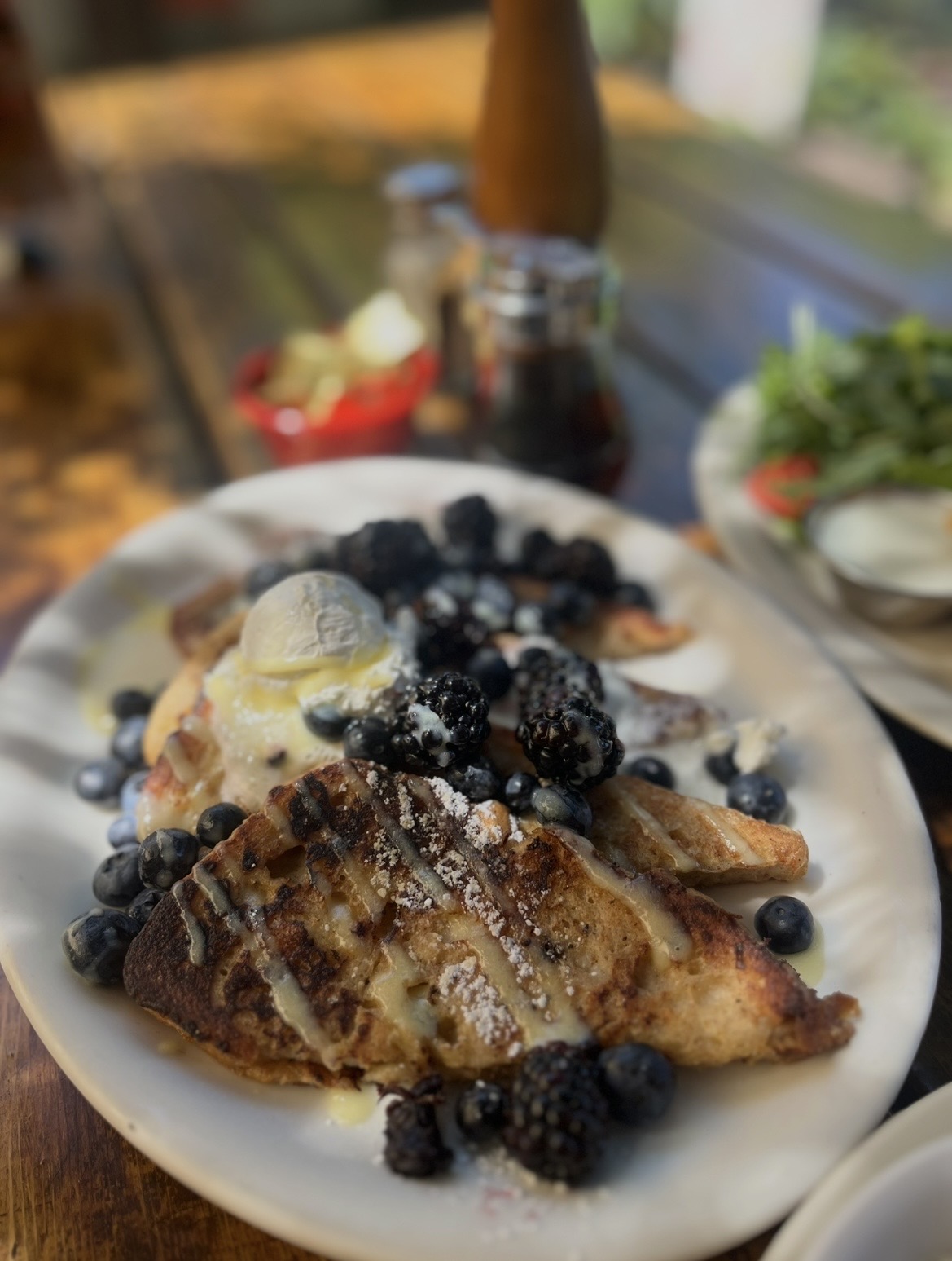 Plate with waffles topped with bananas, blueberries, and powdered sugar, on a wooden table with condiments in the background.