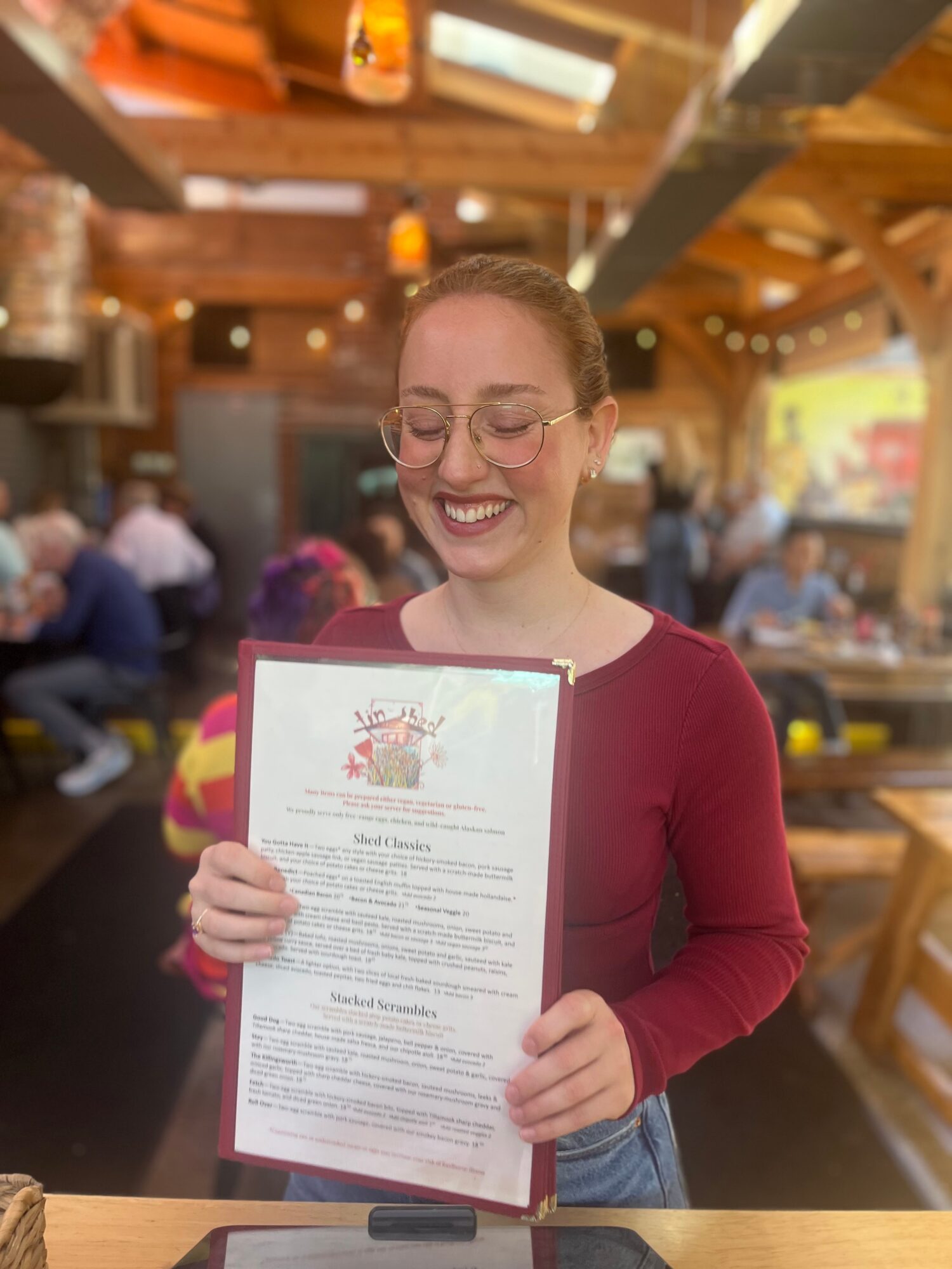 Smiling woman with glasses holding a menu in a lively restaurant with wooden decor and hanging lights.