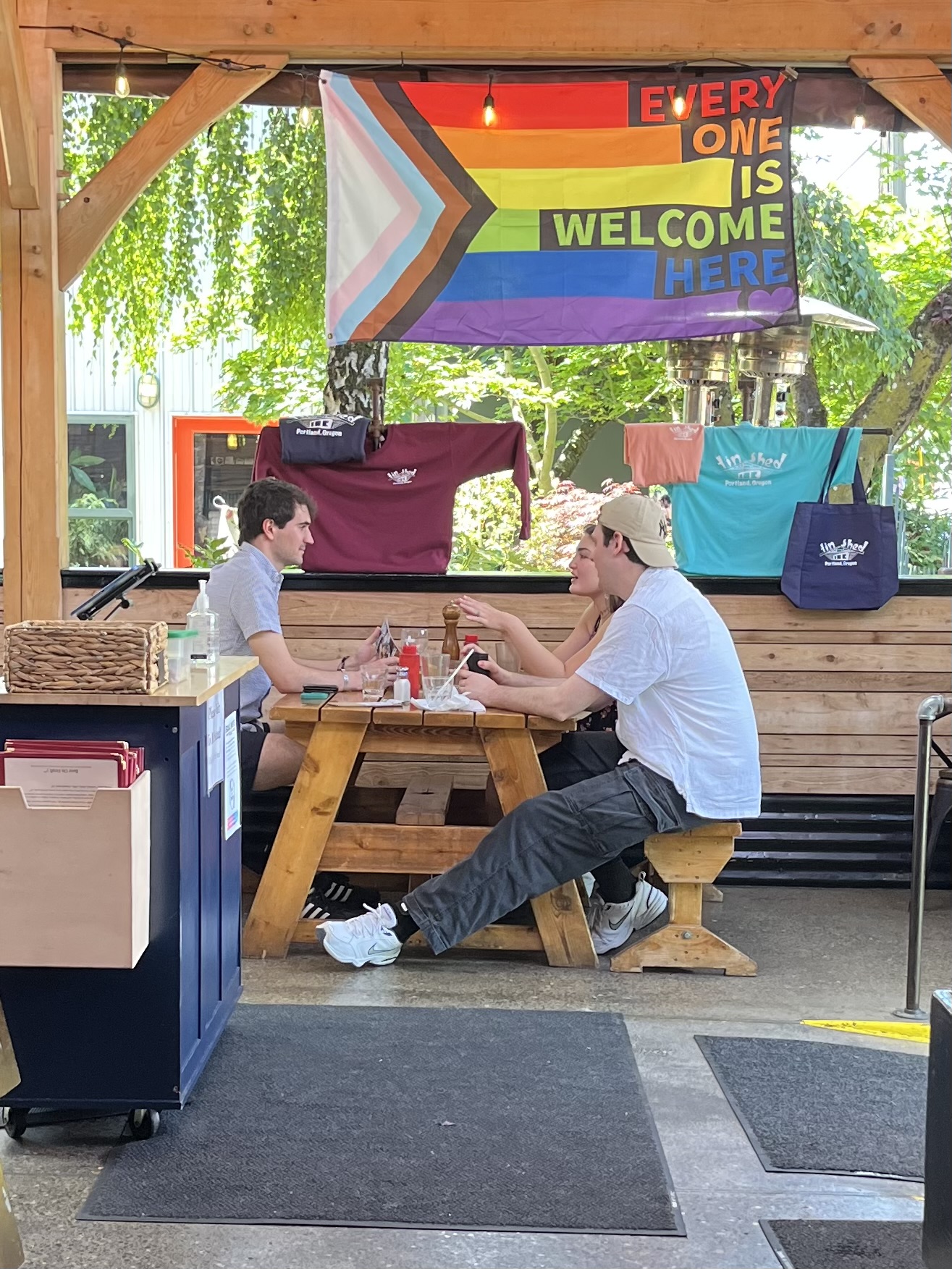 Two people sit at a wooden picnic table outdoors, with rainbow flags and bags hanging behind them, and trees in the background.