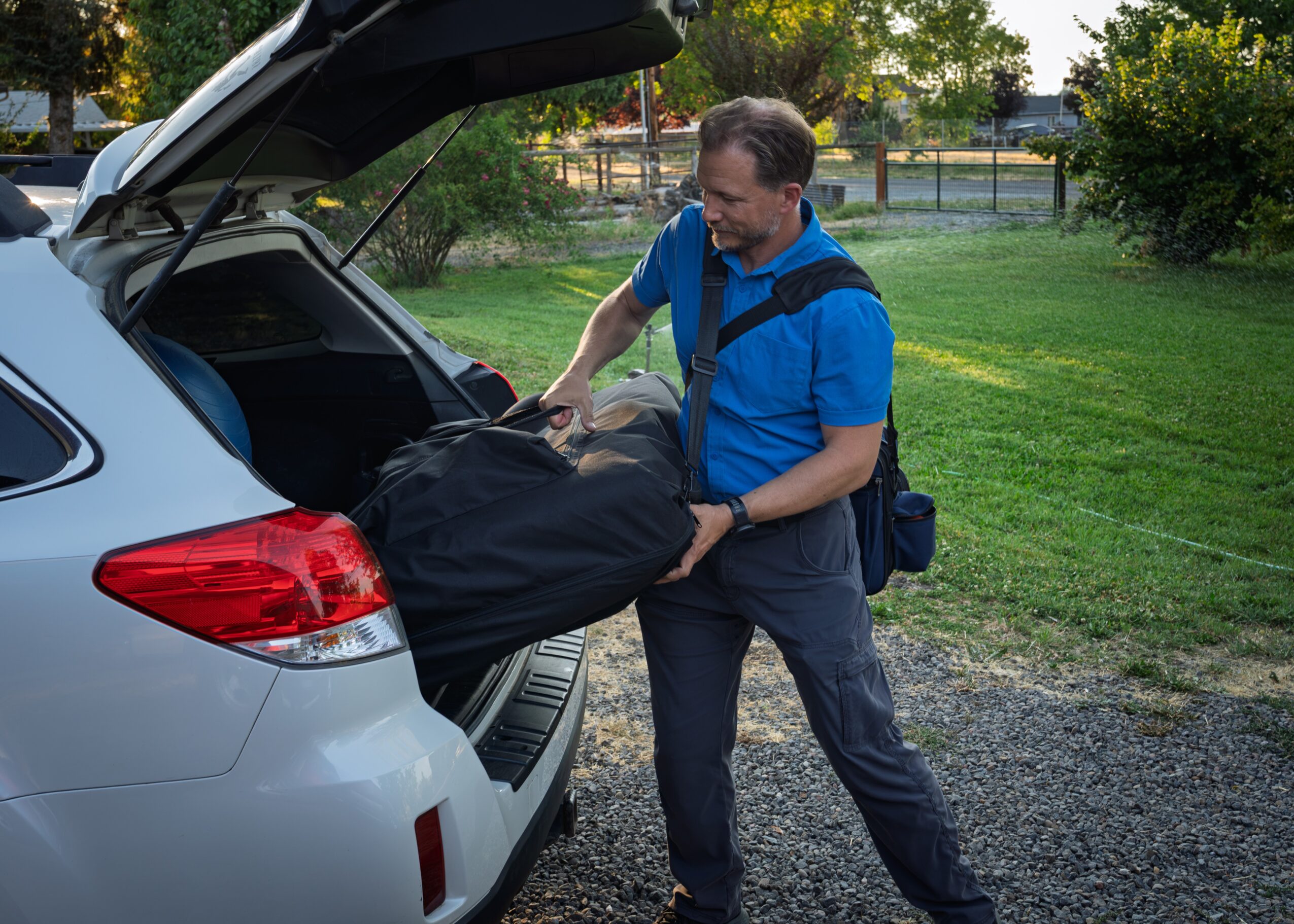 Man loading a large bag into the trunk of a silver car outdoors with trees and a fence in the background.