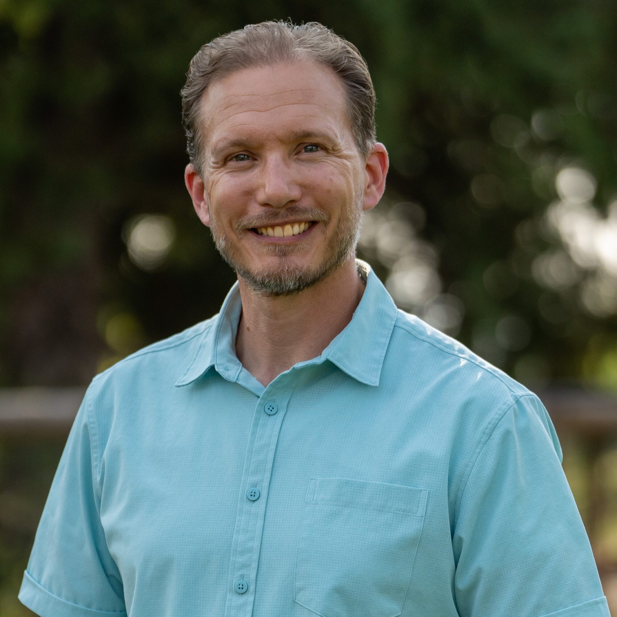 Smiling man with short hair and beard wearing a light blue shirt outdoors with blurred greenery background.