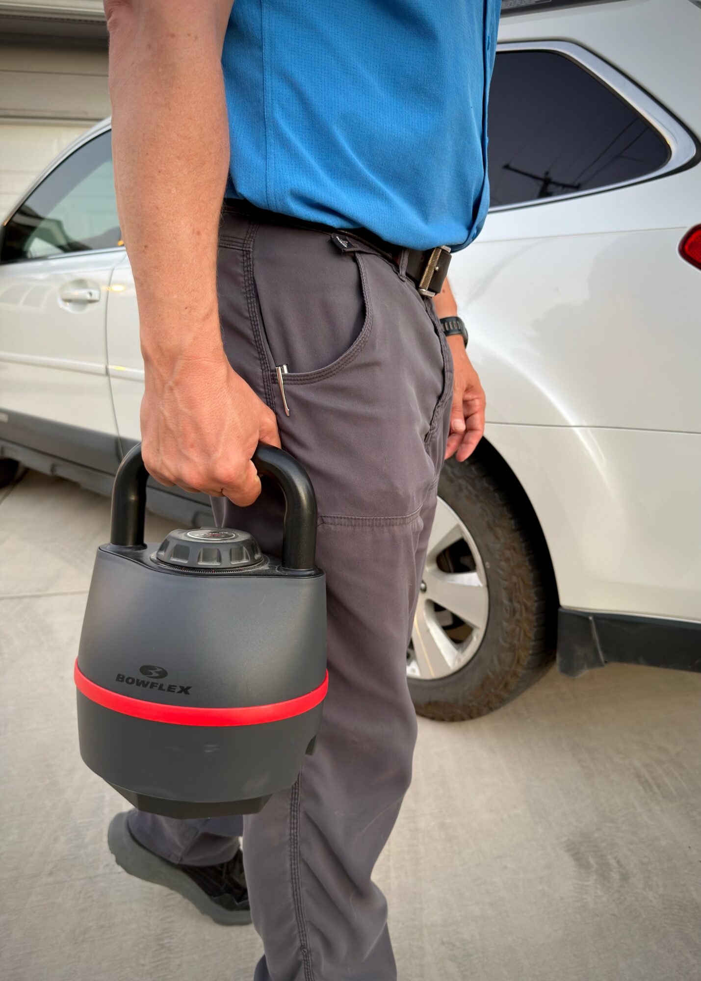 Person holding a portable device in front of a parked car, wearing a blue shirt and gray pants.