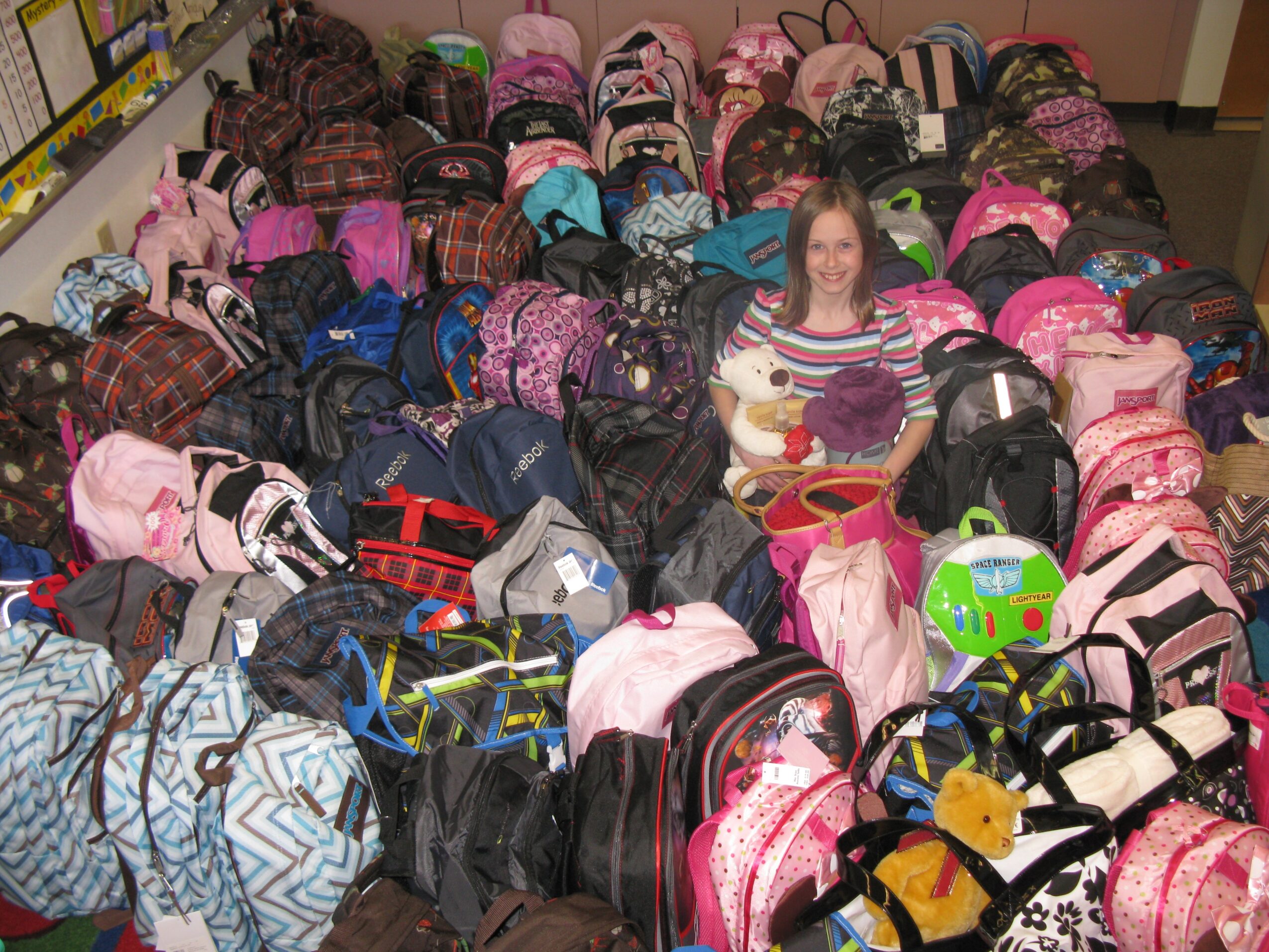 A woman sitting among many backpacks and bags in a store, holding a stuffed animal and smiling.