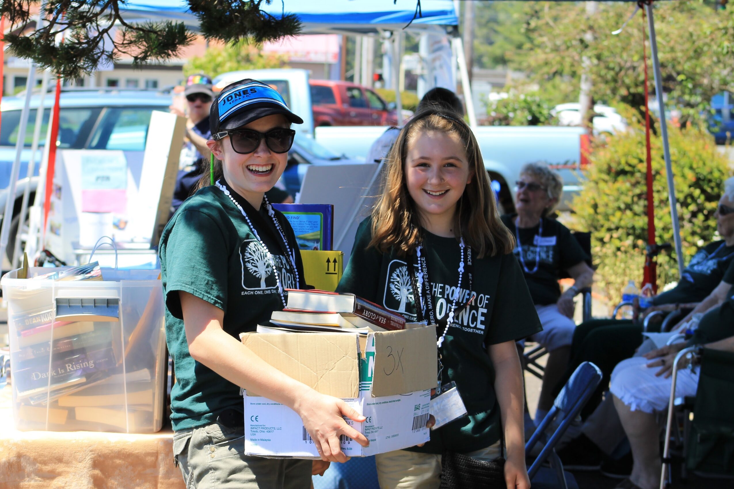 Two smiling young women holding boxes and packages outdoors at an event, with tents and people in the background.