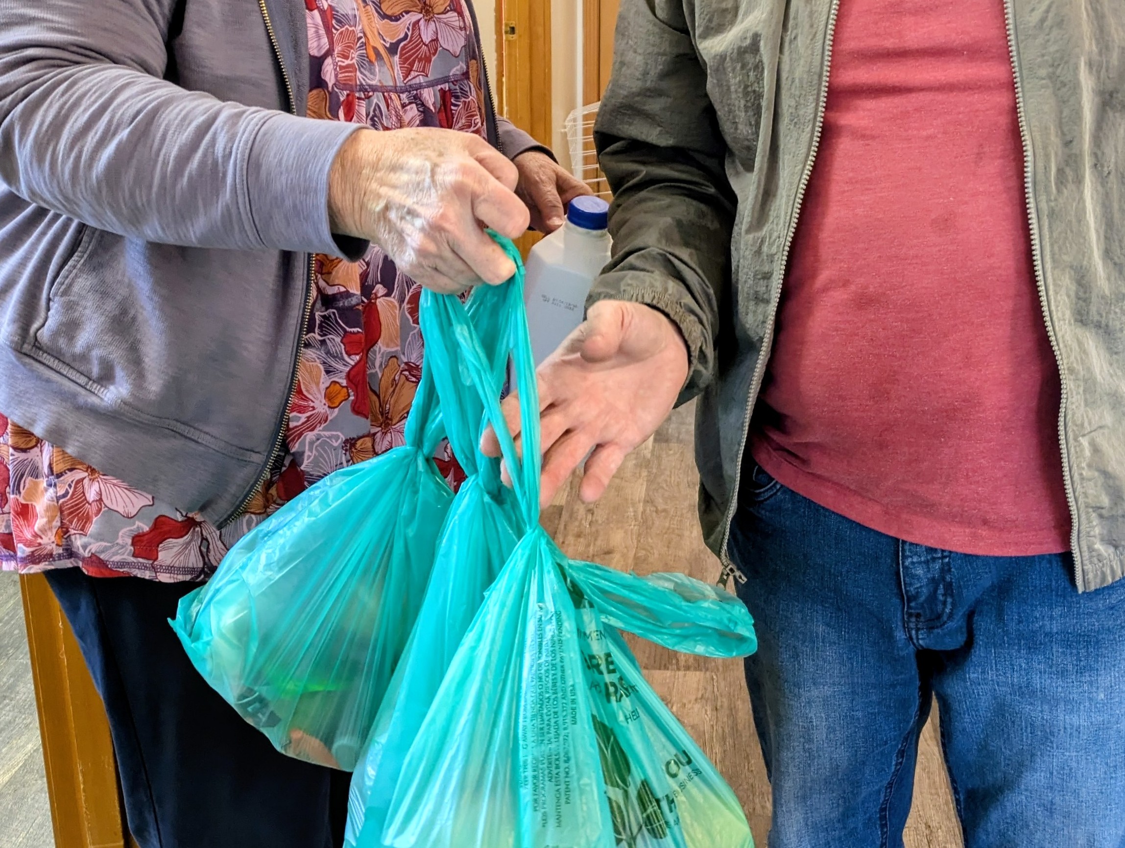 Two people exchange a blue plastic bag, one holding a bottle, in a room with wooden paneling.
