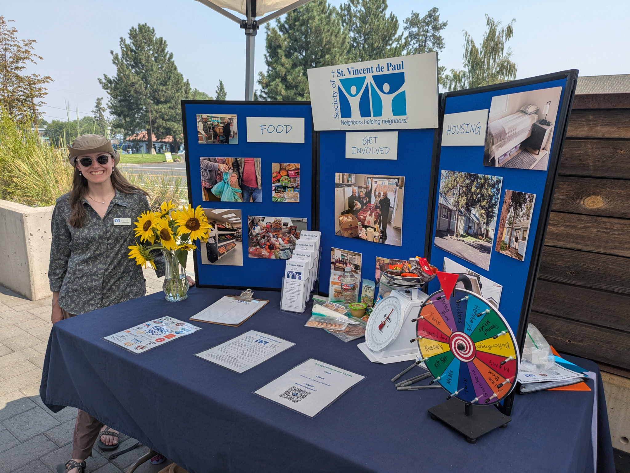 Woman standing next to a display booth with photos, a table with papers, a colorful wheel, and sunflowers.