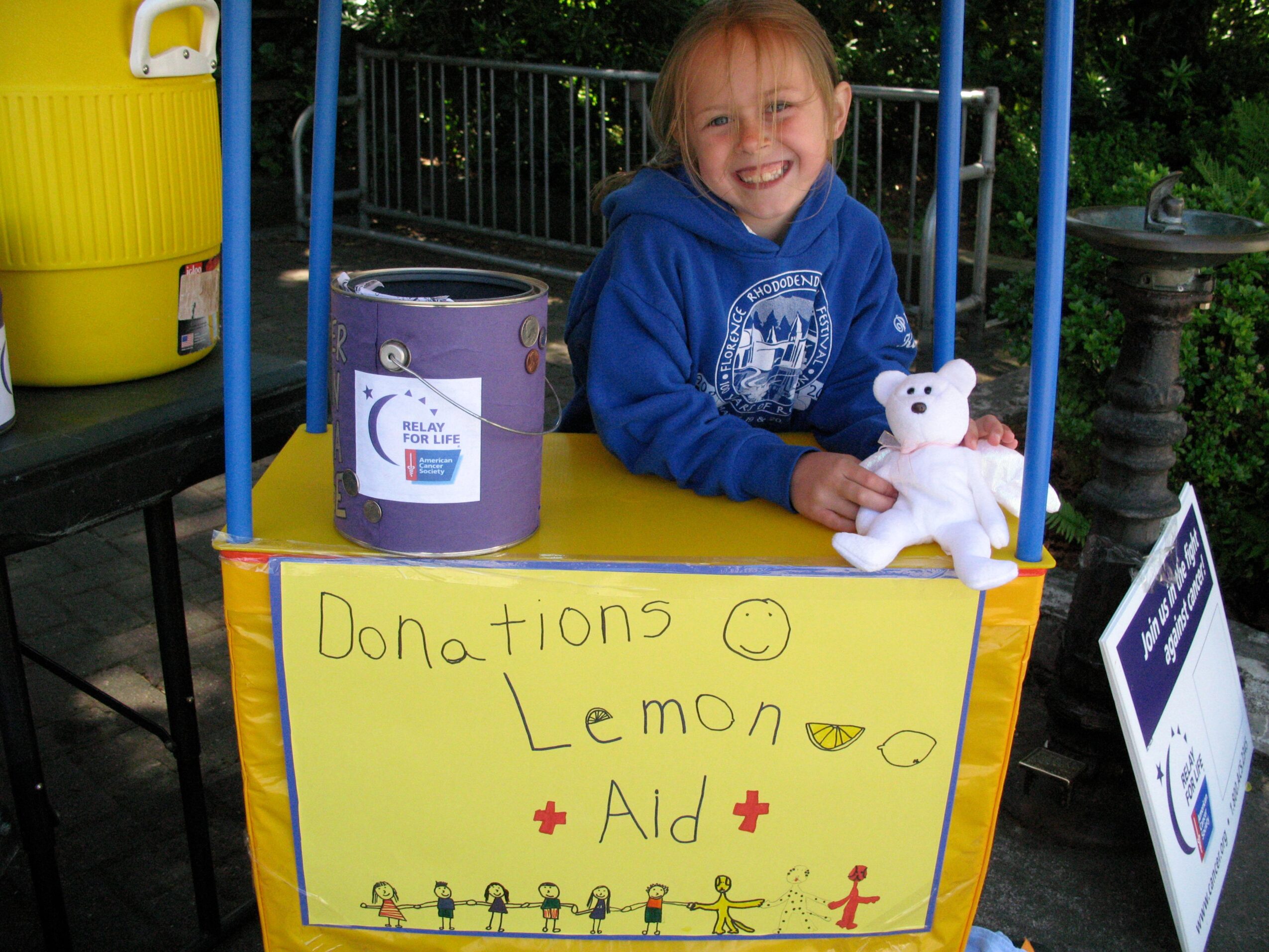 Child smiling on a yellow table holding a teddy bear, with a donation sign and outdoor background.