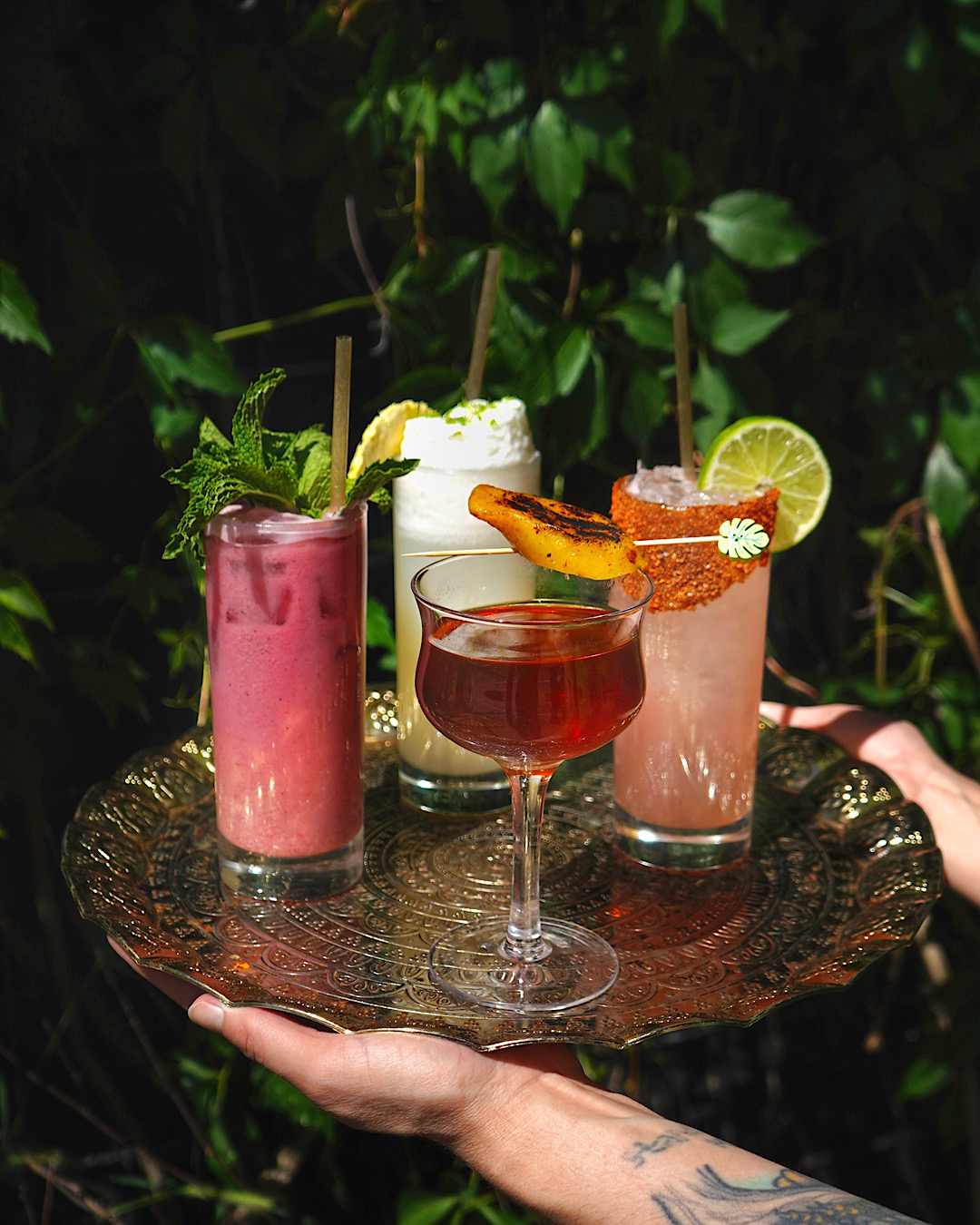 Four colorful cocktails on a decorative tray held by a person with a tattooed hand, against a leafy background.