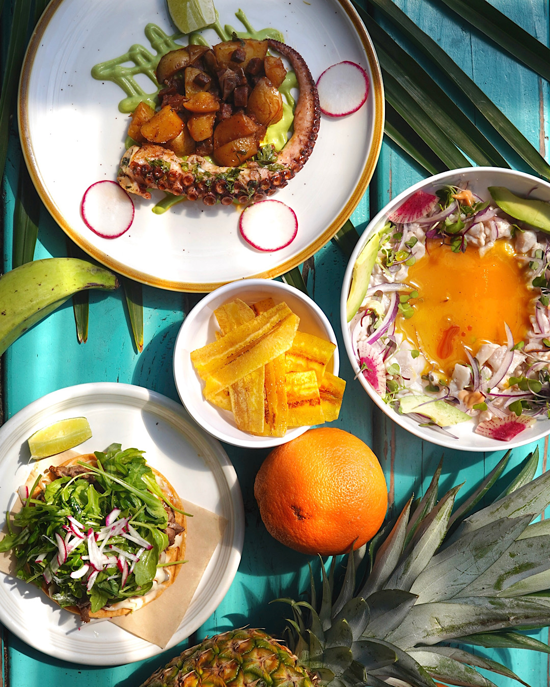 Plate with meat and vegetables, bowl of sliced plantains, salad with greens and radishes, orange, and a bowl of soup with an egg yolk, on a turquoise surface.