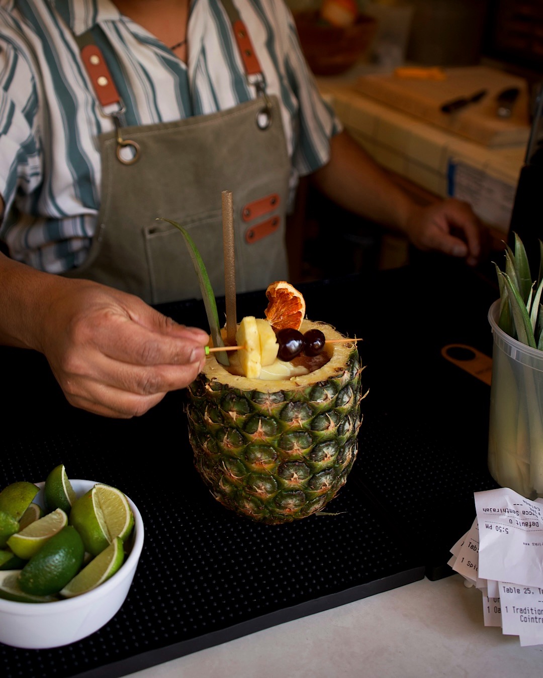 Person preparing a fruit cocktail inside a hollowed pineapple, garnished with various fruits and a green straw, on a black surface.