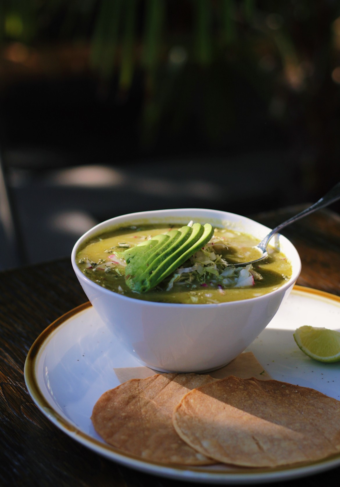 Bowl of green soup with avocado slices and a spoon, served with flatbread and lime wedge on a white plate.