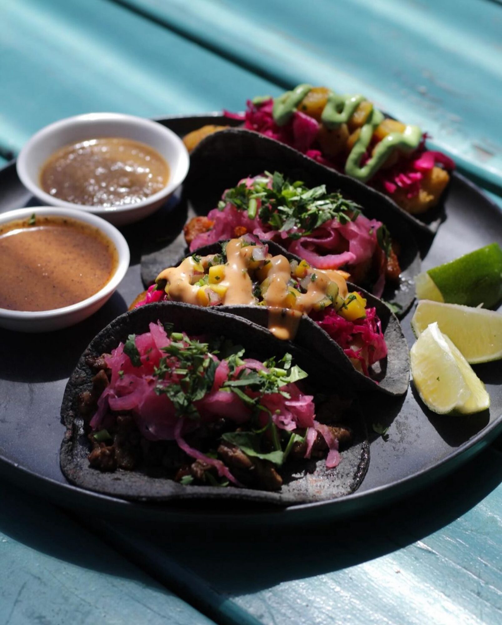 Three tacos with colorful toppings, two small bowls of sauce, and lime wedges on a black plate.