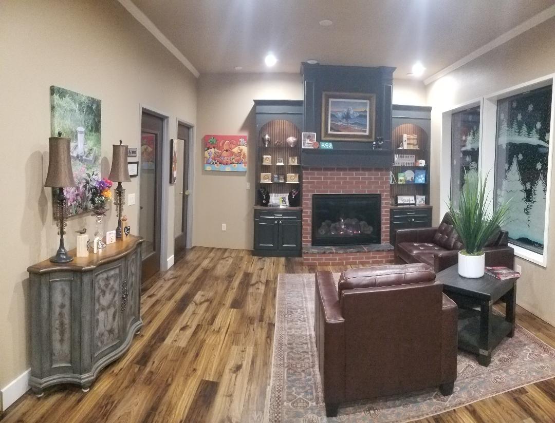 Living room with fireplace, two armchairs, large window, and wooden floor, decorated with paintings and plants.
