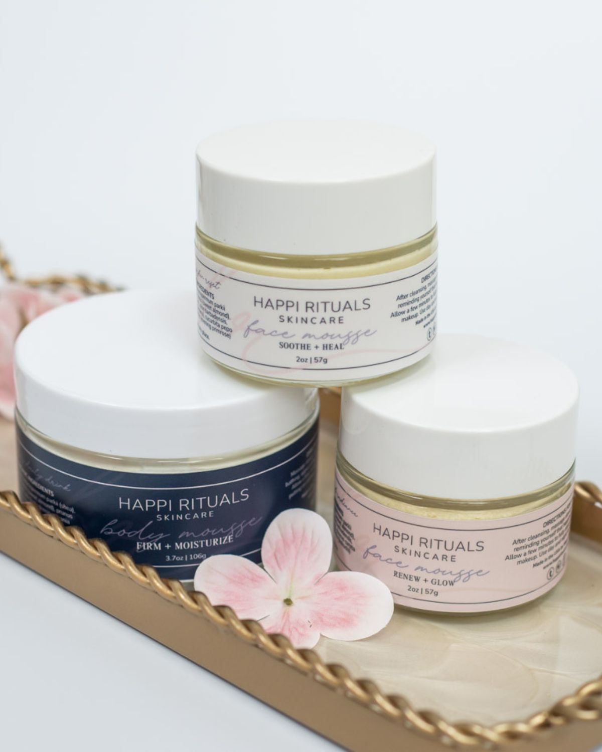 Three skincare jars on a tray with pink flower petals, two jars are white and one is dark blue, all labeled 'Happi Rituals'.